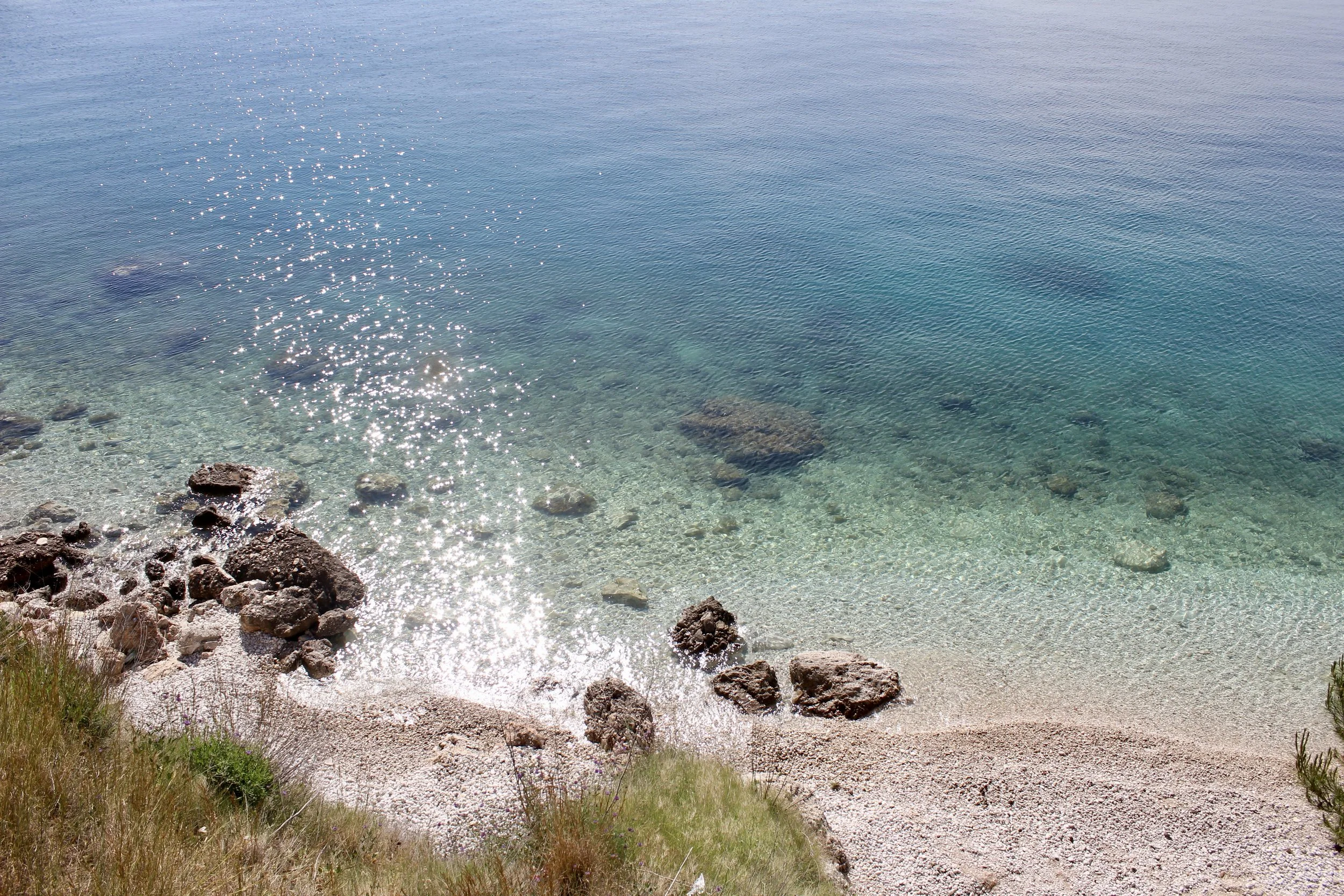 Clear turquoise ocean water with rocks along the shoreline, grassy and pebble-covered beach, bright sunny day.