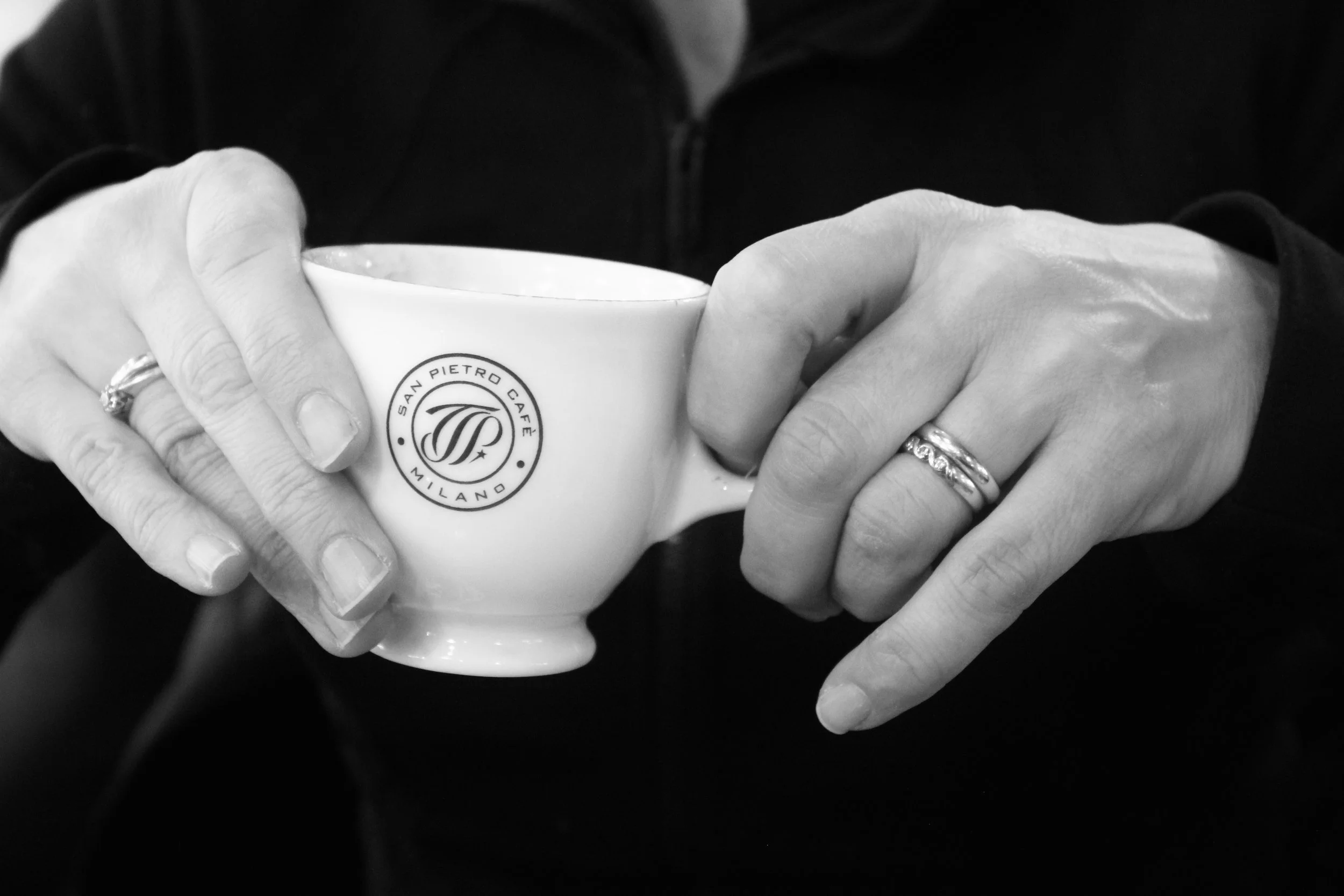 Black and white photograph of two hands holding a white coffee cup. The hands have rings and are positioned close together. The cup has a logo that says 'San Pietro Café Milano'.