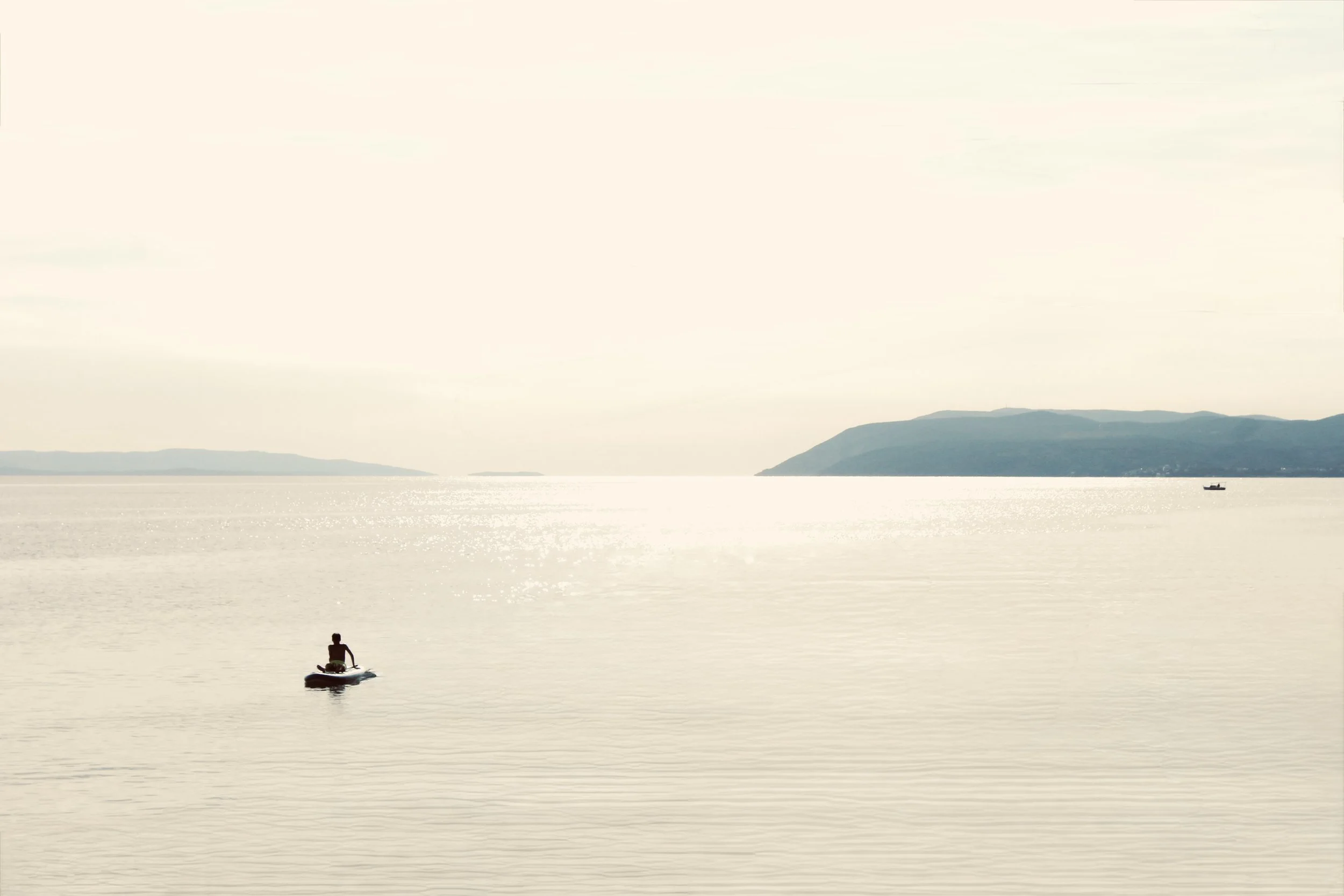 A person paddleboarding on a calm body of water with distant hills and a boat in the background, under a pale sky.