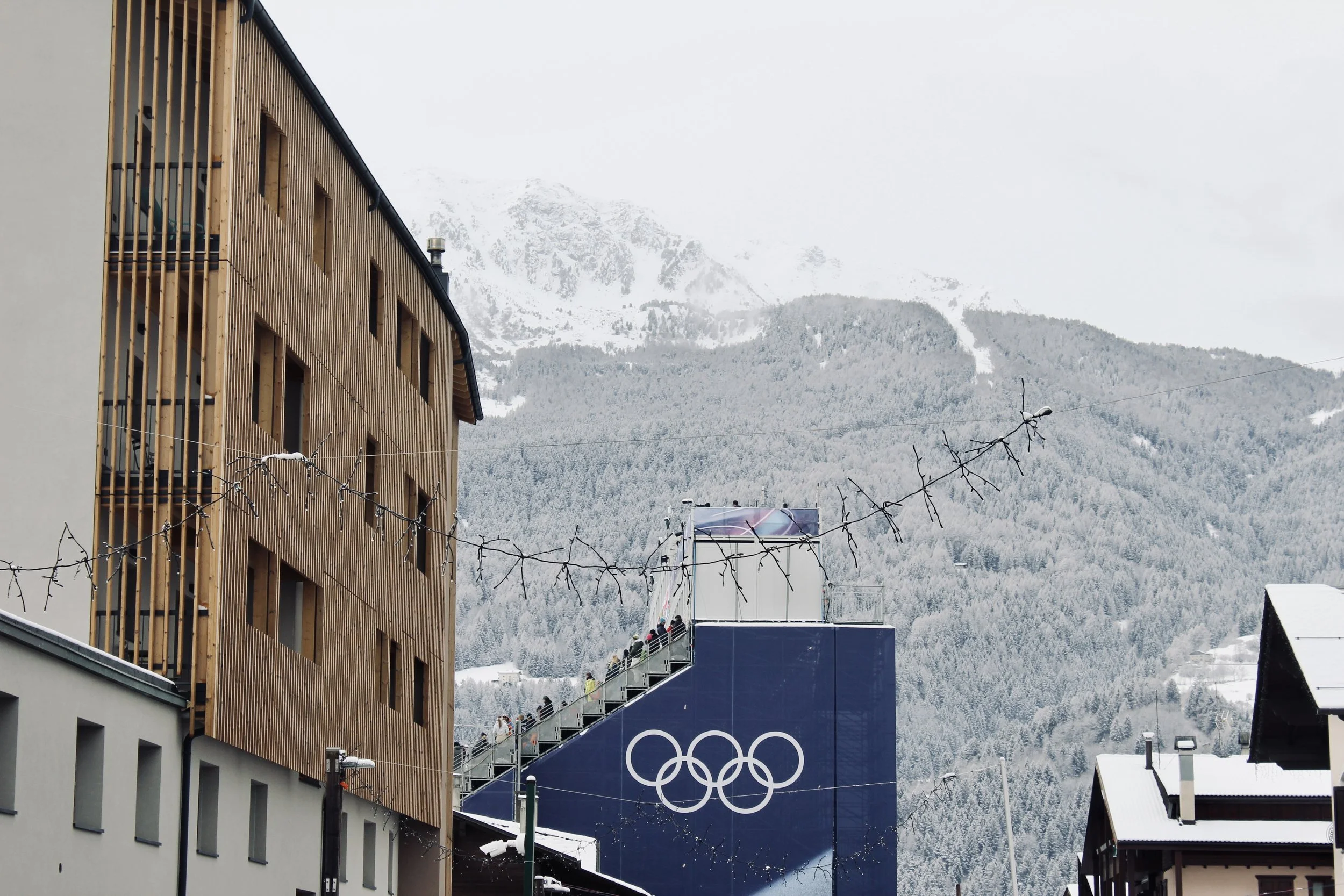 A building under construction with wooden exterior is in the foreground, and snow-covered mountains with trees and ski slopes are in the background. There are people on a suspended walkway near the building, and an Olympics banner featuring five inte