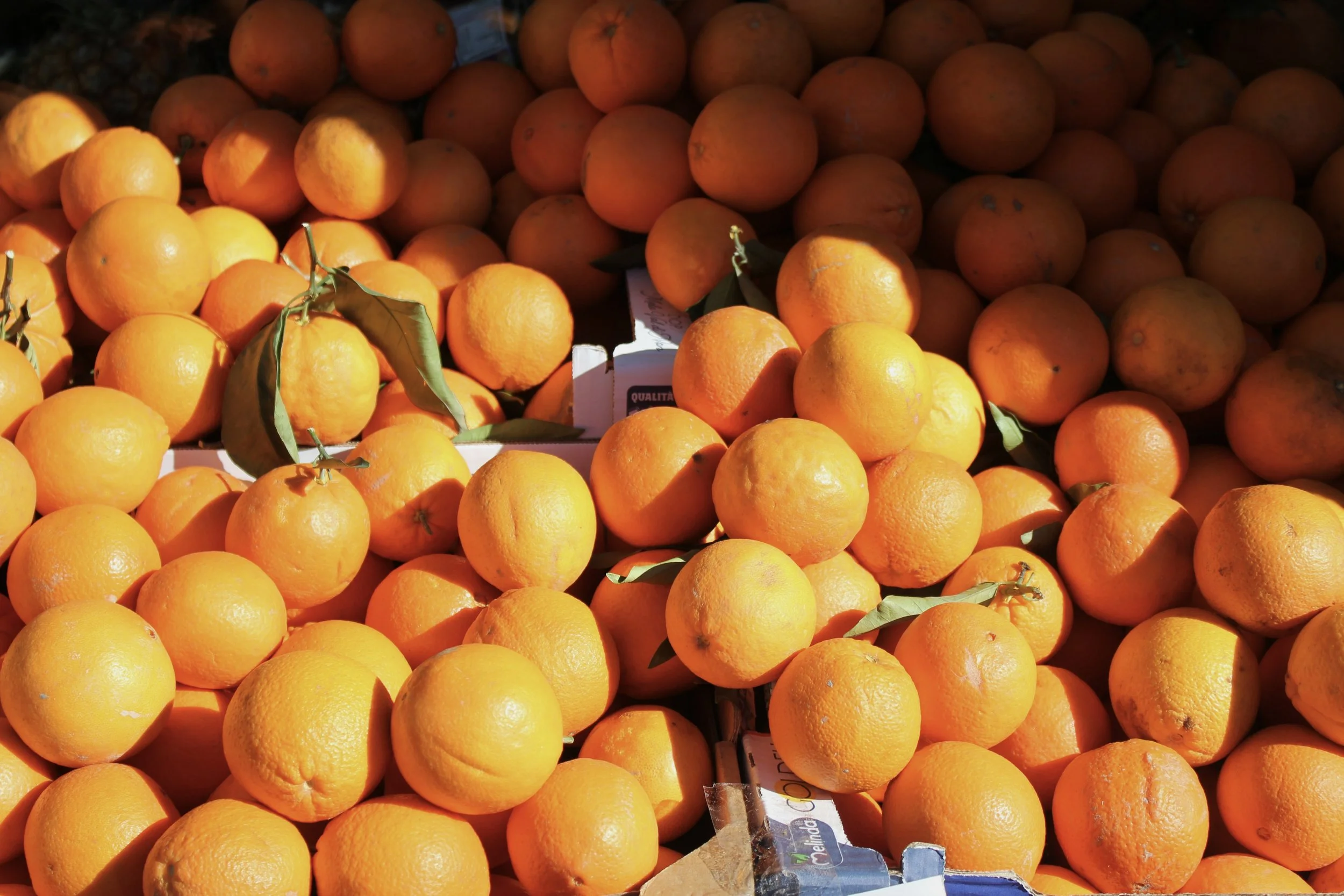 A large pile of fresh oranges, some with green leaves still attached, displayed at an outdoor market or grocery store.