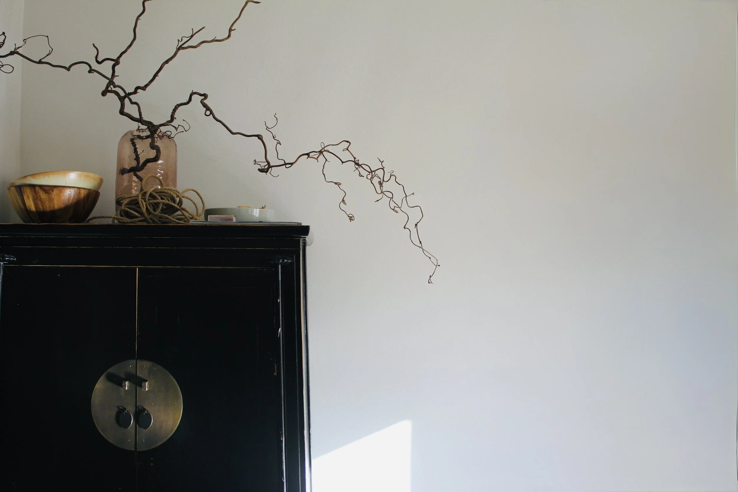 A black display cabinet with a circular metallic lock, topped with a large ceramic bowl, a decorative glass jar with twigs, a ball of twine, and a small white dish with a yellow item, against a plain white wall.