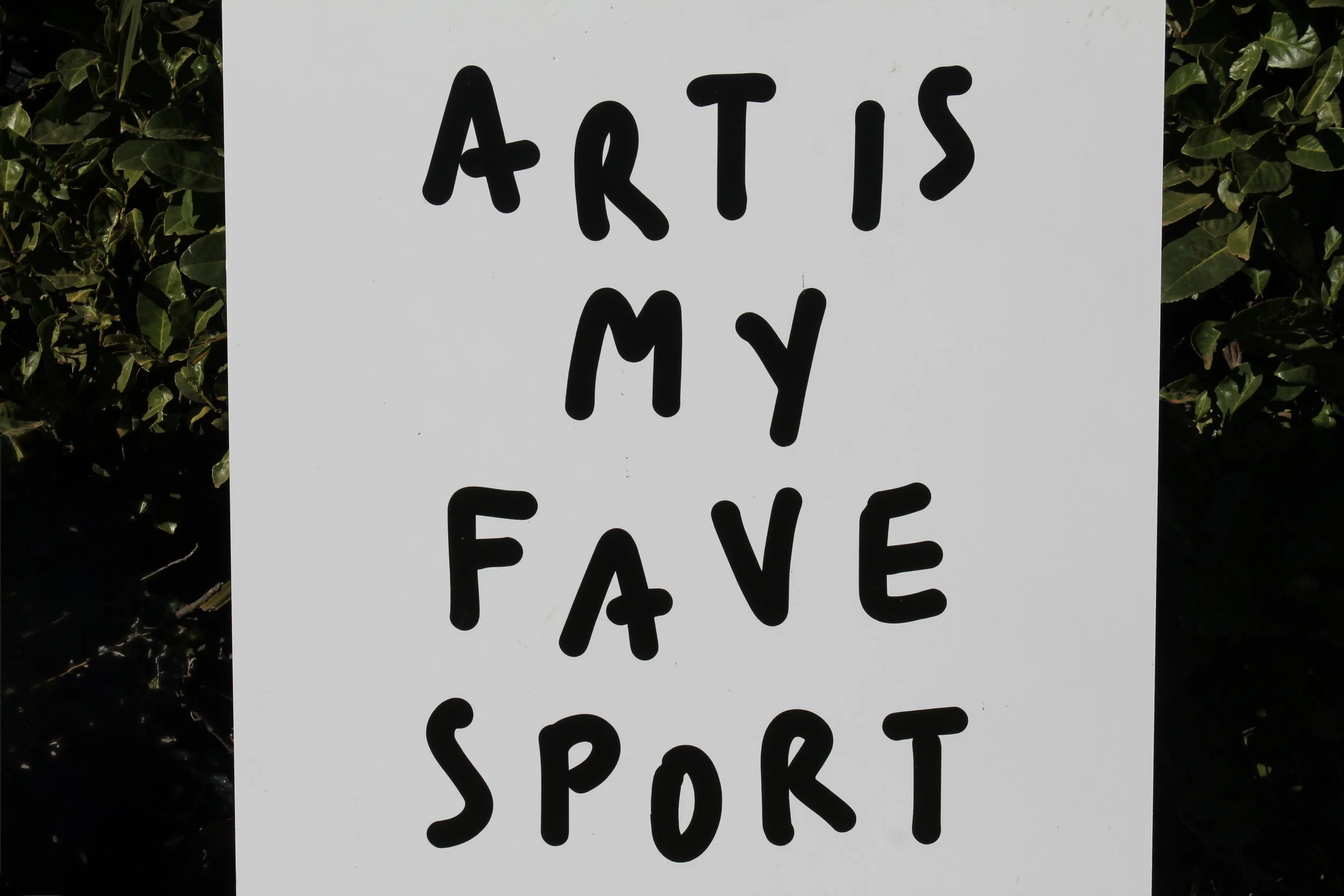 A white sign with black handwritten text reading 'ARTIS MY FREE SPORT', with green bushes or leaves in the background.