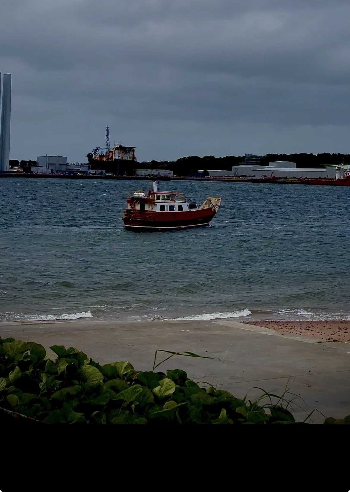 A small red and white boat floating on the water near the beach, with industrial buildings and a large ship in the background under a cloudy sky.