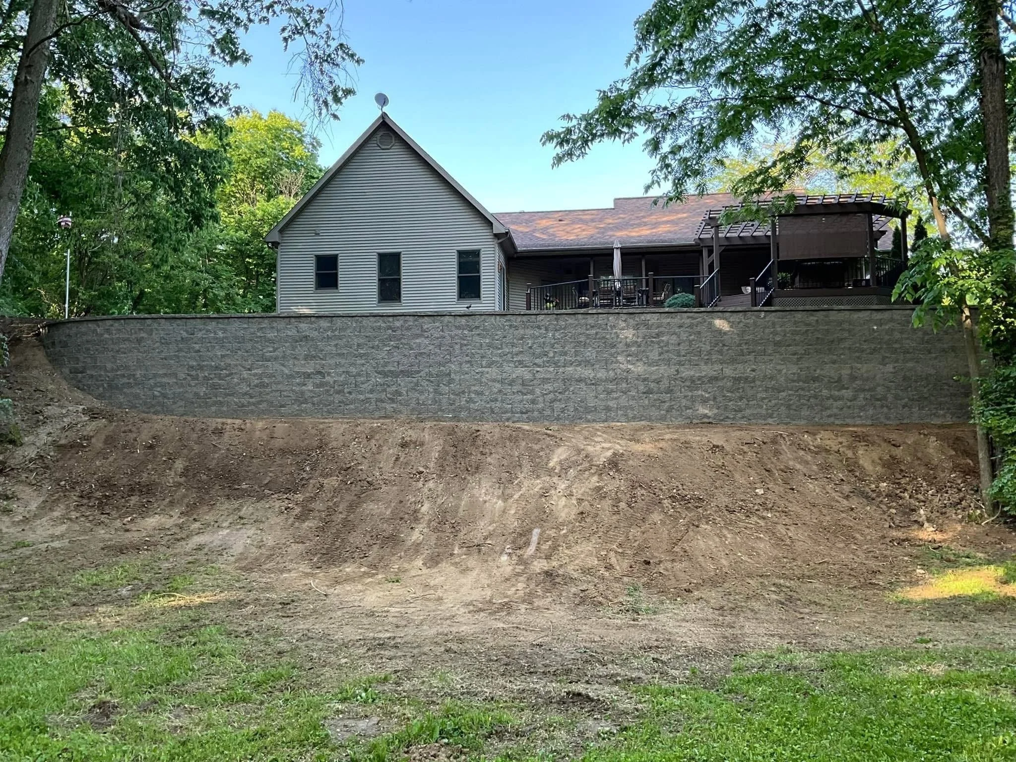 View of a house with a sloped roof and gray siding, surrounded by green trees, with a stone retaining wall in the foreground and an open backyard patio area with outdoor furniture.