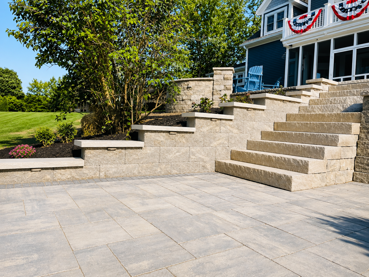 Stone patio with steps leading up to a house with a porch decorated with red, white, and blue bunting, and a yard with green grass, trees, and flower beds.