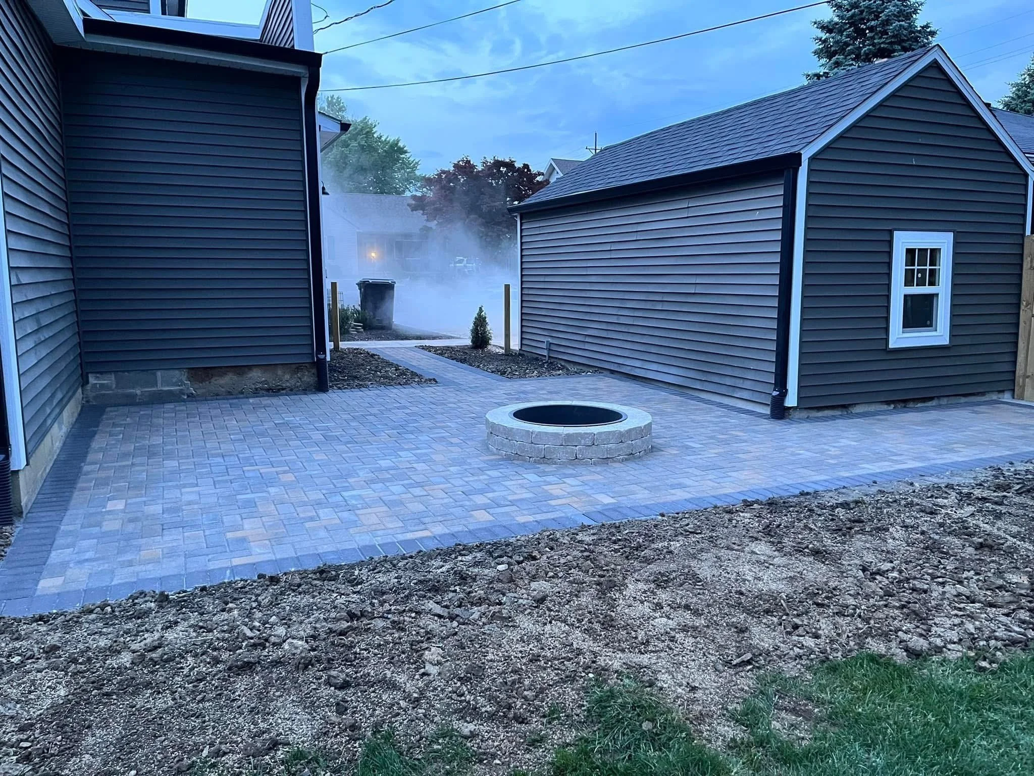 An outdoor patio area with newly installed brick pavers, a round fire pit, and a small shrub. Two houses with siding and shingle roofs are visible, along with a tree and a neighboring house in the background. Smoke or steam is emanating from behind a