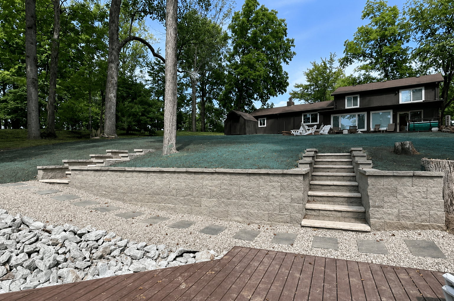 Backyard with stone retaining wall, stairs, and a deck, featuring a house with patio furniture and surrounded by trees.