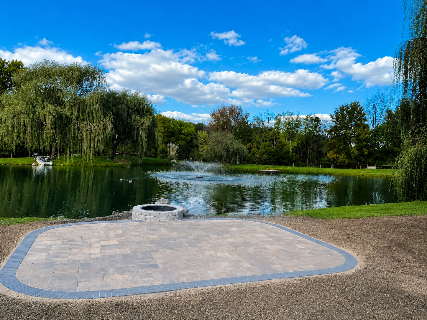 A peaceful park with a pond surrounded by trees under a blue sky with white clouds. A fountain is in the middle of the pond, and a paved area with a fire pit is in the foreground.
