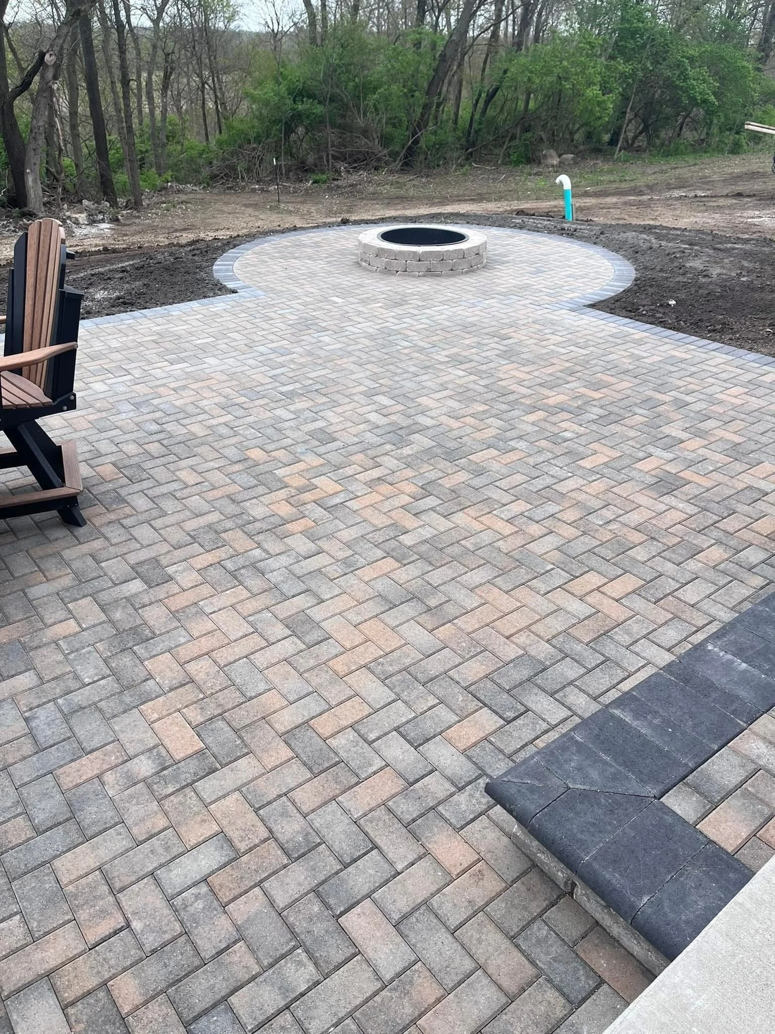 A paved backyard patio area with a fire pit and a wooden bench, surrounded by dirt, with trees and bushes in the background.