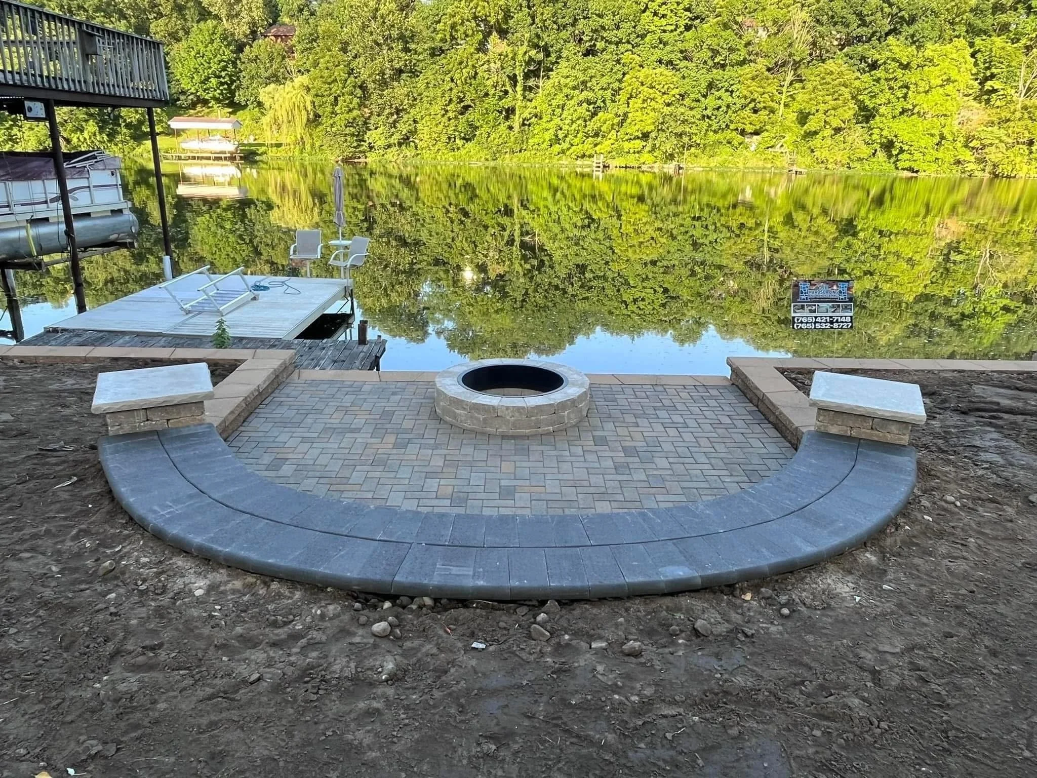 Backyard with a patio, fire pit, chairs, and a view of a lake surrounded by trees, with some boat docks and a sign with phone numbers.