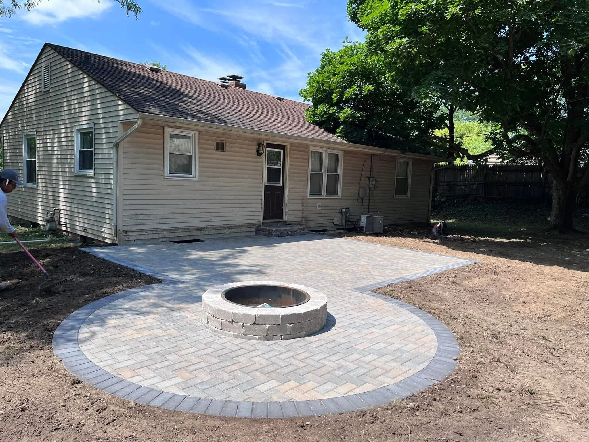 Backyard with newly paved circular patio and fire pit, with house, trees, and clear blue sky in the background.