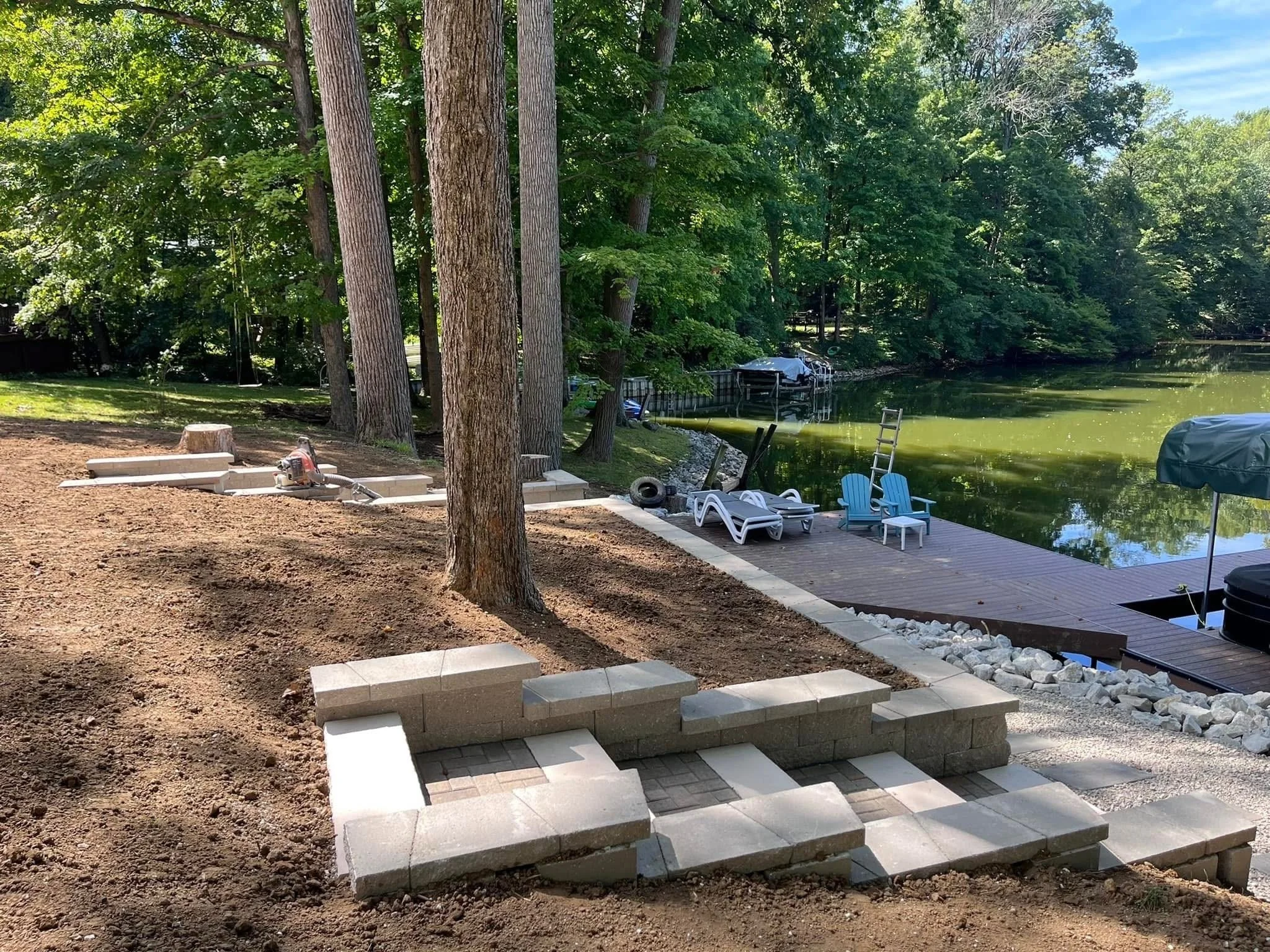 Backyard scene with trees, a pond, and a deck with lounge chairs, some stones, and a ladder by the water during daytime.