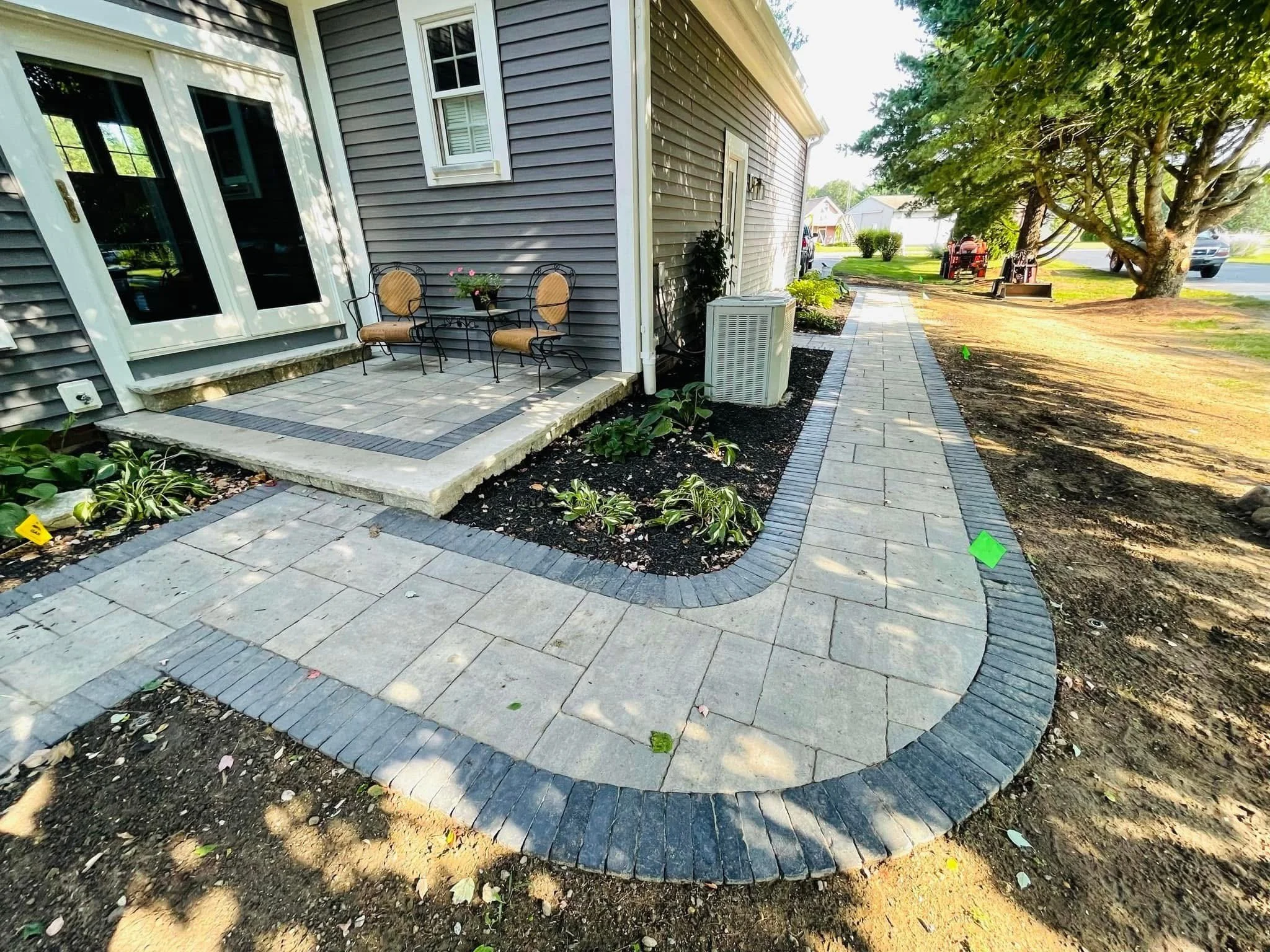 The image shows a freshly paved brick patio area next to a house with gray siding. There are two chairs and a small table on the patio, and surrounding flower beds and a newly laid walkway. Trees and houses are visible in the background on a sunny da