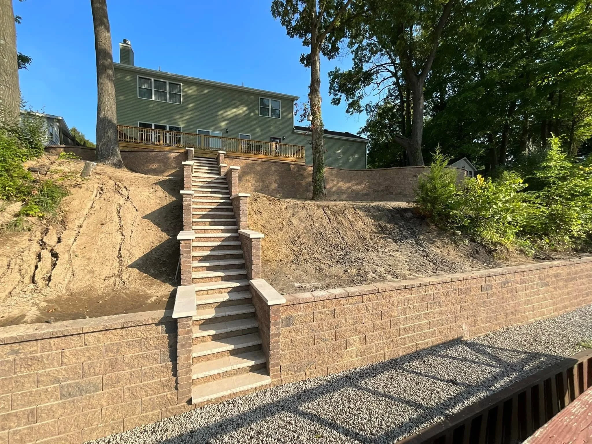 View of the backyard backyard showing a house on a hill with a wooden deck, several trees, and a brick staircase with steps leading up to the deck, with a stone wall and gravel in the foreground.