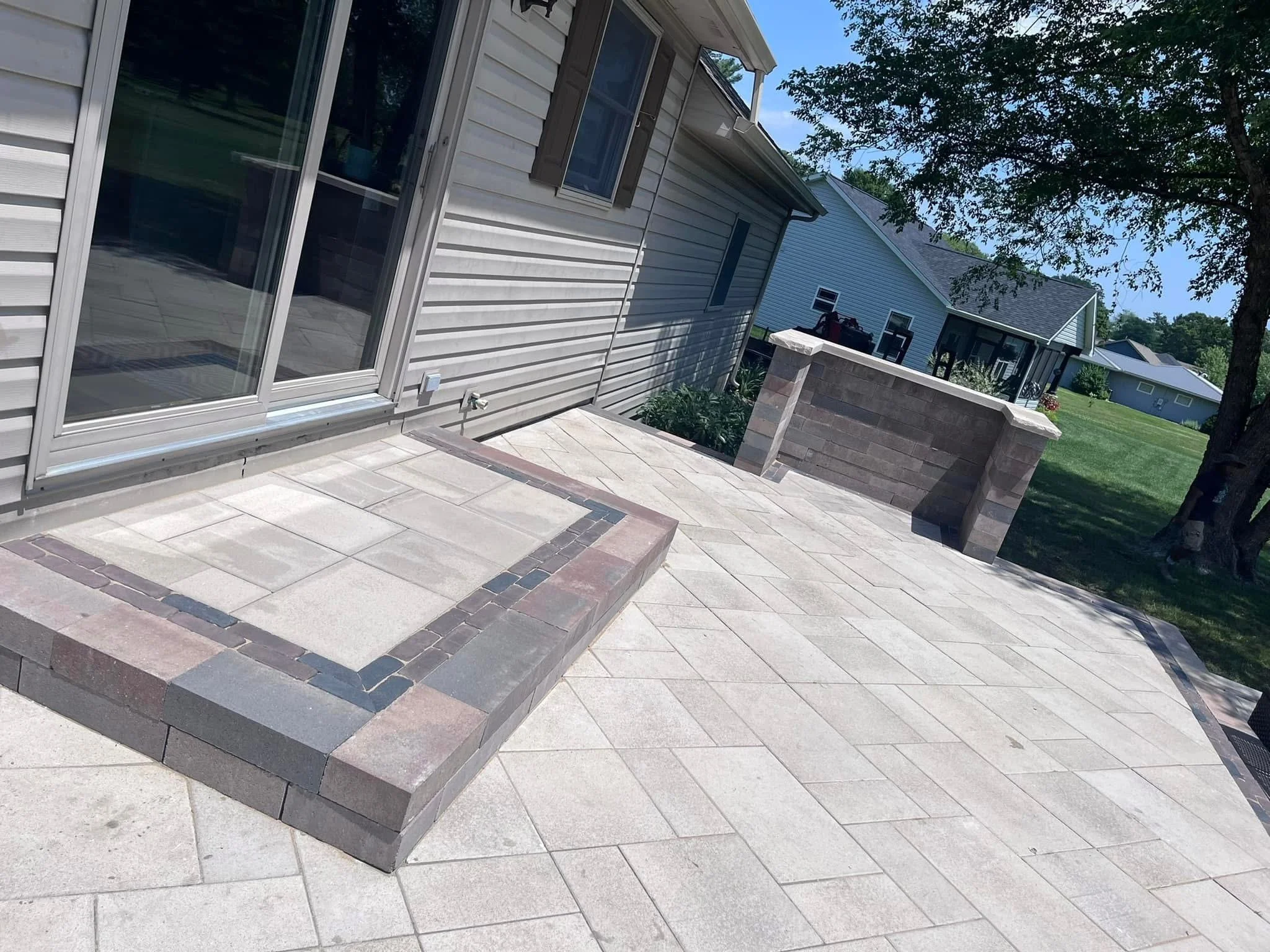 A residential backyard with a sliding glass door, tiled patio, stone steps, a low brick wall, green lawn, trees, and neighboring houses.