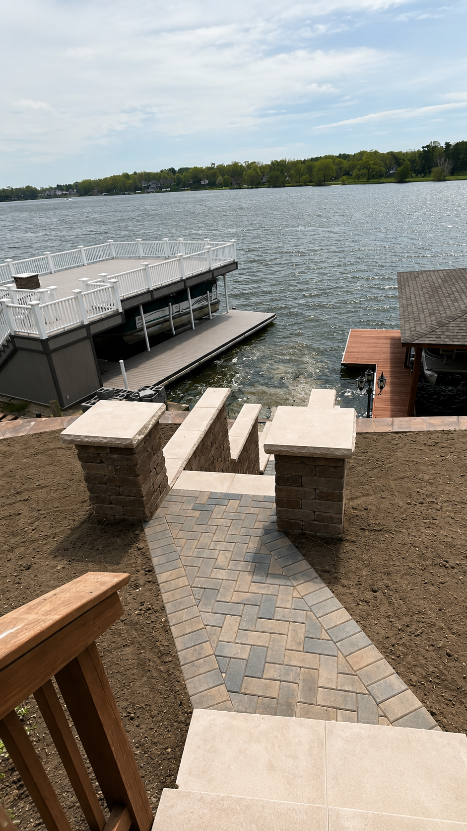 Pathway leading down to a waterfront with a boat dock and a pontoon boat, surrounded by a patio and brick steps, overlooking a lake with trees on the opposite shore.