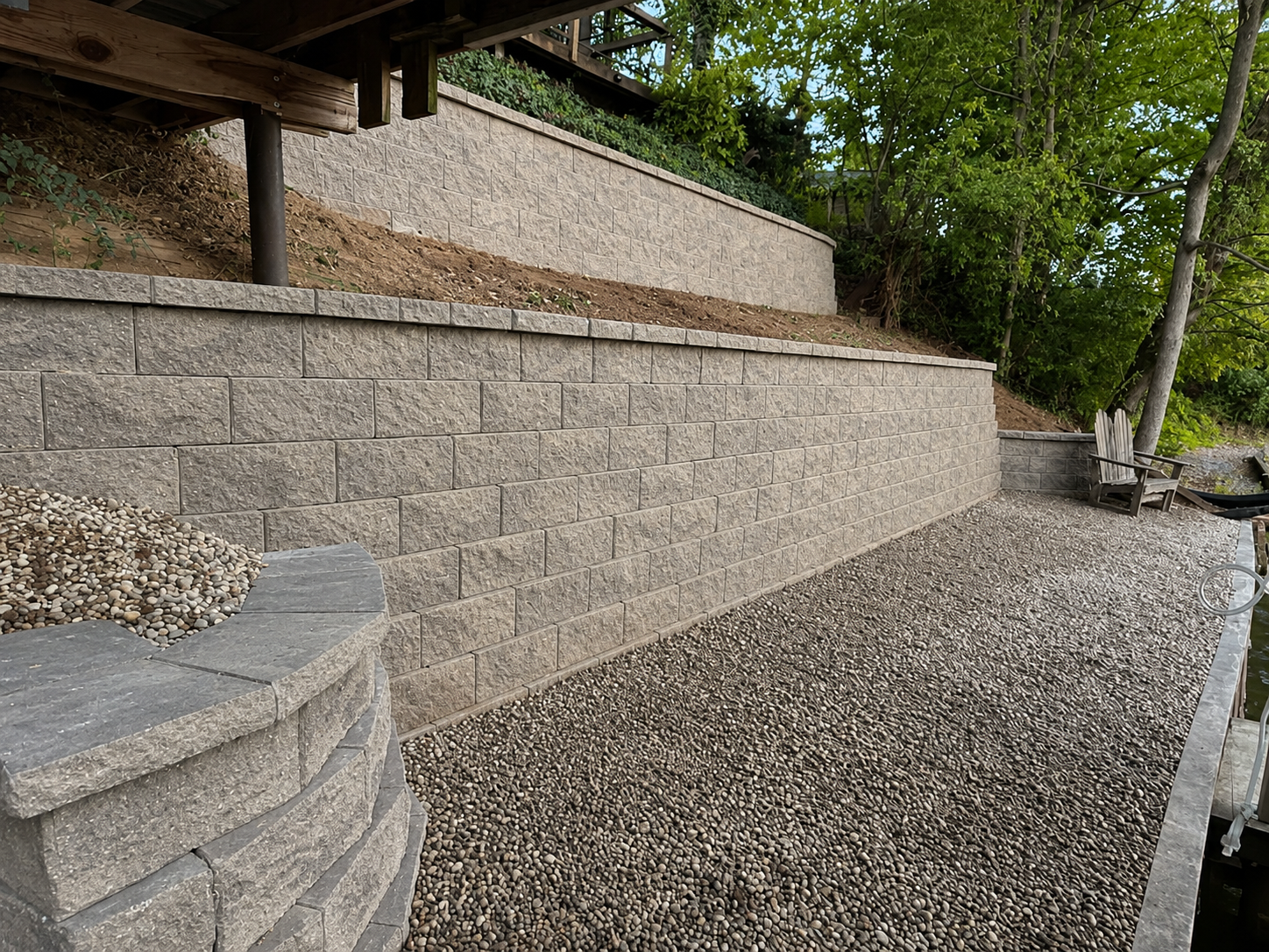 A backyard with a multi-tiered gray retaining wall, gravel ground cover, and a wooden bench near a small body of water surrounded by trees.