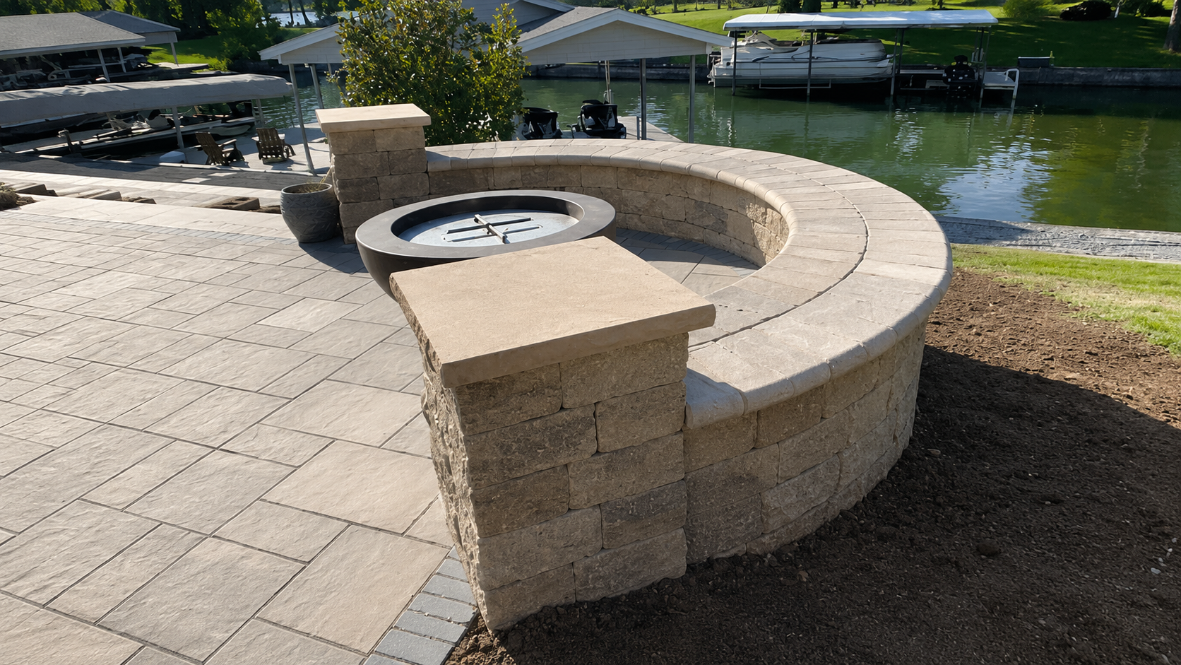 A waterfront patio with a stone firepit, a curved stone bench, and outdoor seating by a canal with boats. There are covered boat docks and green grass in the background, with clear weather and sunlight.
