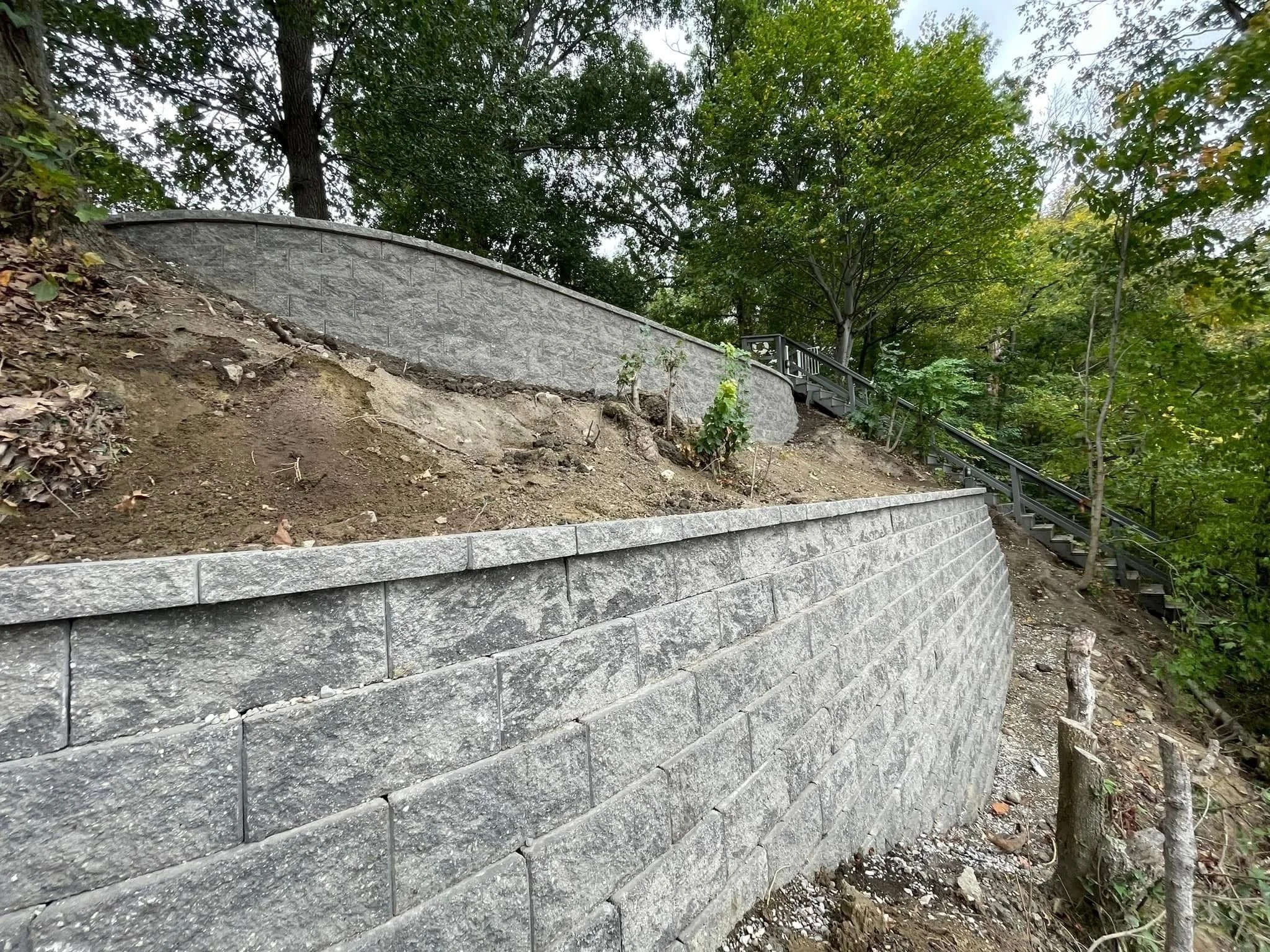 A stone retaining wall with a staircase and metal railing, set in a wooded area, with open dirt and soil in the foreground.