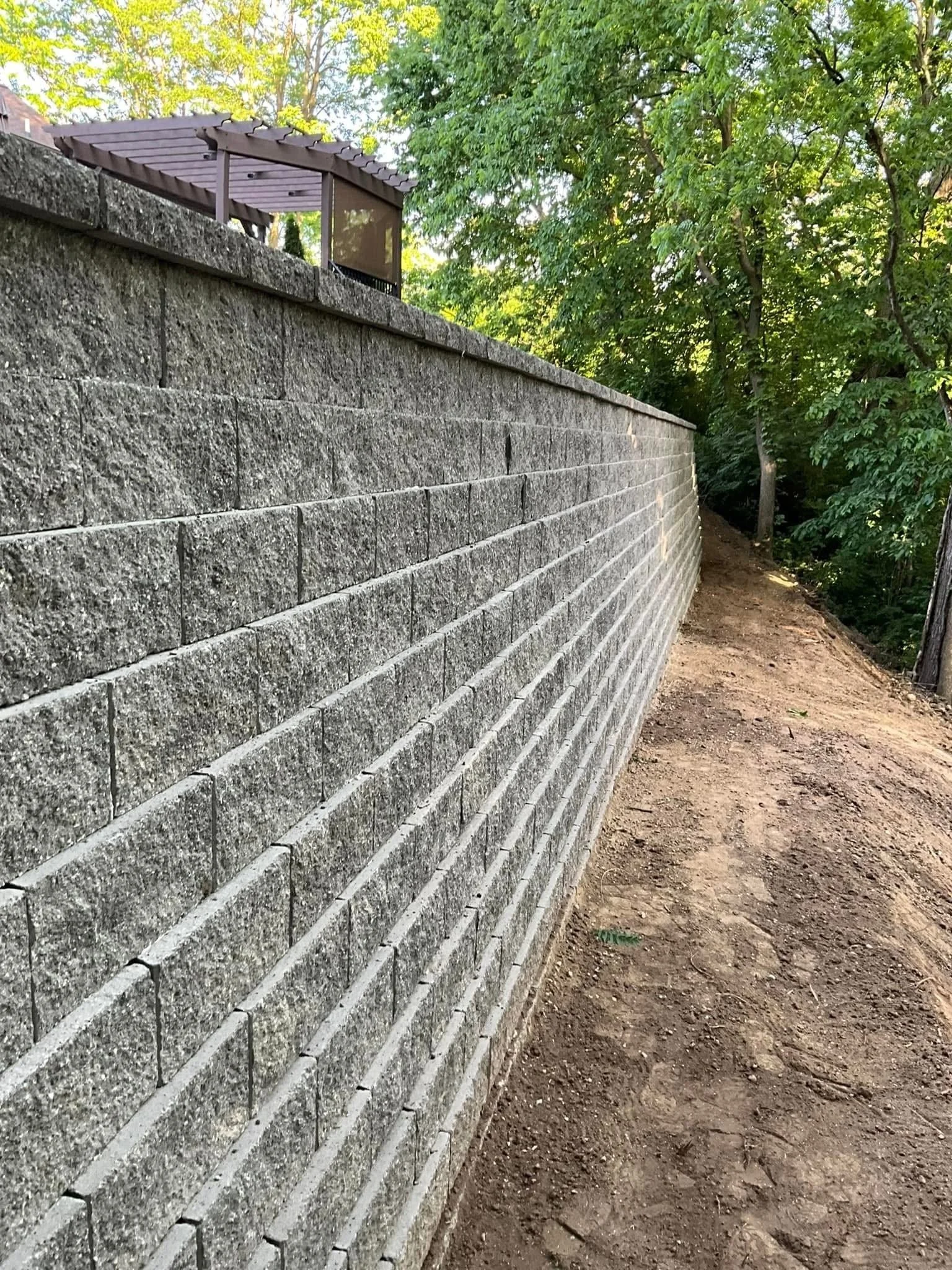 A stone block retaining wall with a wooden fence on top, running along a dirt pathway surrounded by green trees.