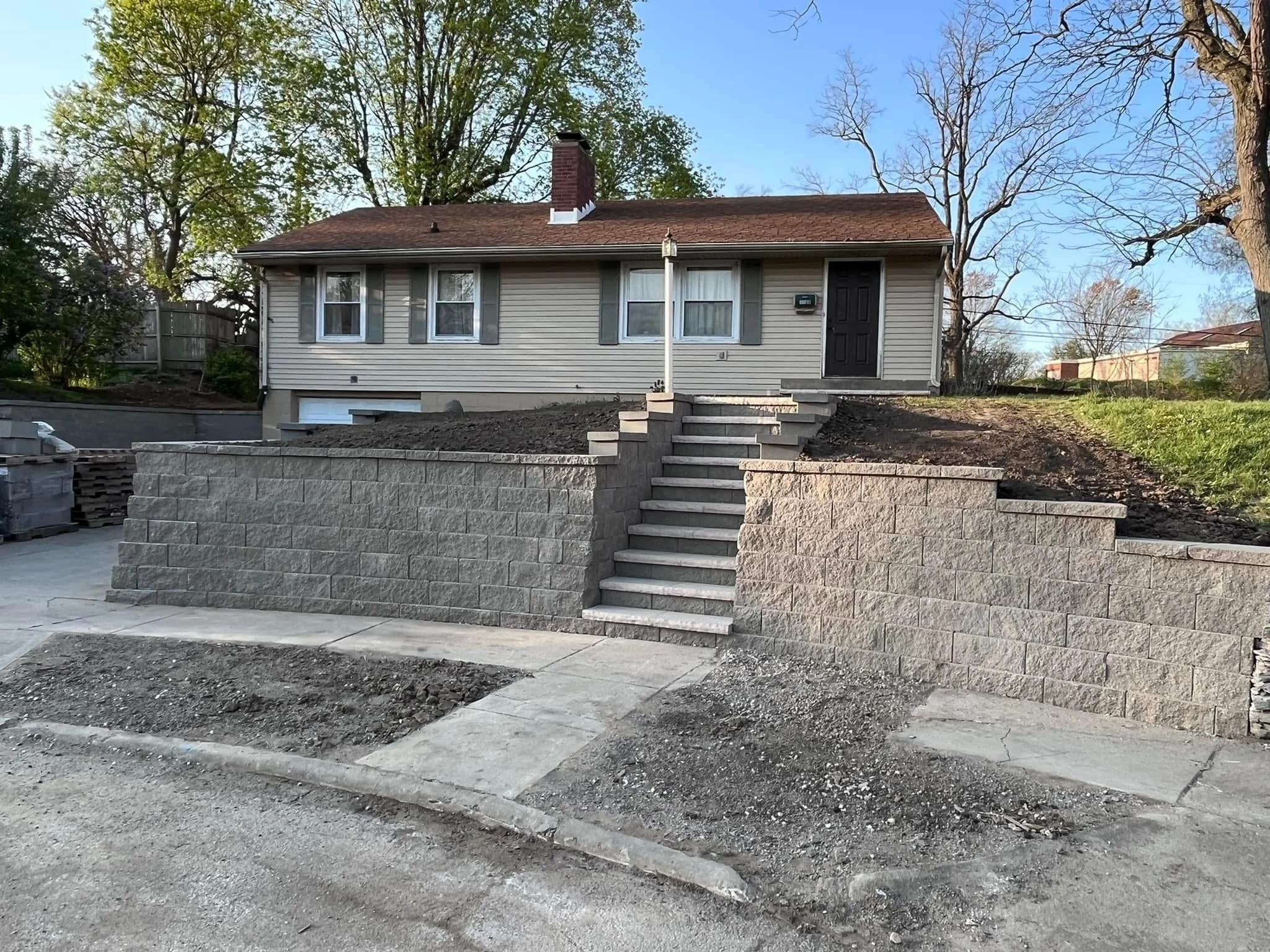 A house under construction or renovation with an unfinished front yard. The house is beige with gray shutters and a black door. There are concrete stairs leading up to the door, but the ground around the stairs is still being worked on. The sky is cl