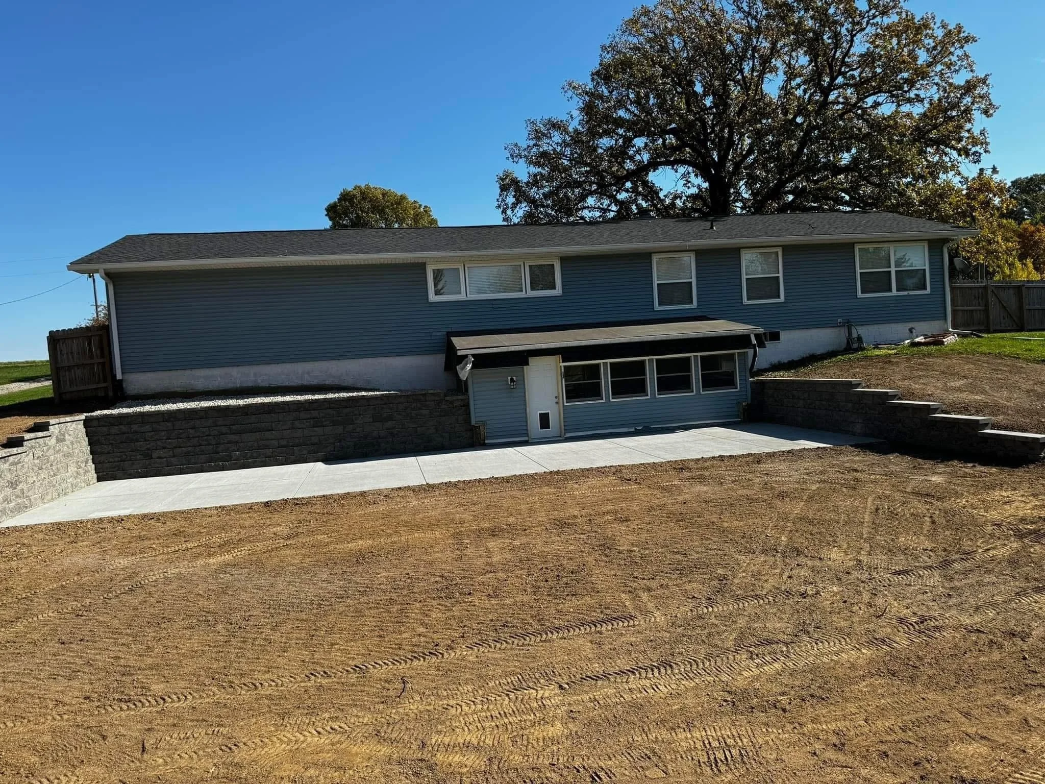 A house with a blue exterior, multiple windows, and a concrete driveway, with a large tree and a clear blue sky in the background.