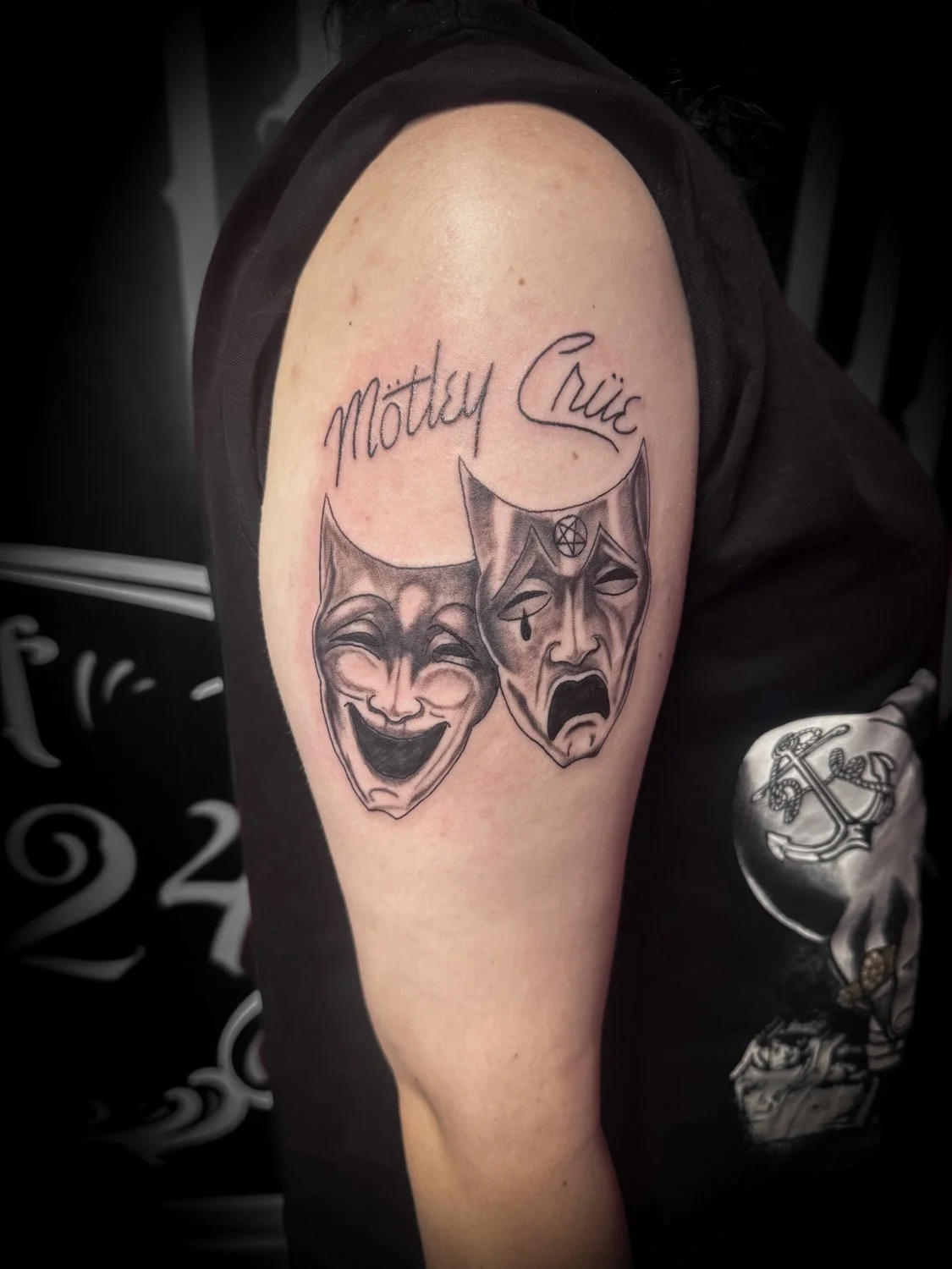 Tattoo on person's upper arm featuring two theatrical comedy and tragedy masks, with the name "Mötley Crüe" written above in cursive.