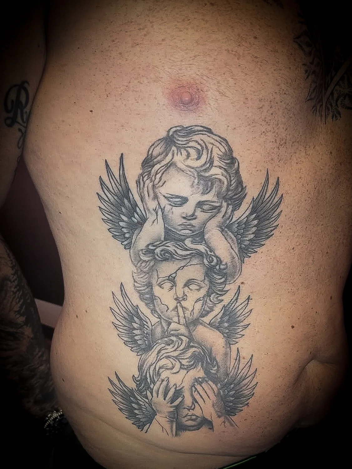 Tattoo of three angelic faces with wings, each face expressing different emotions, on a person's chest.