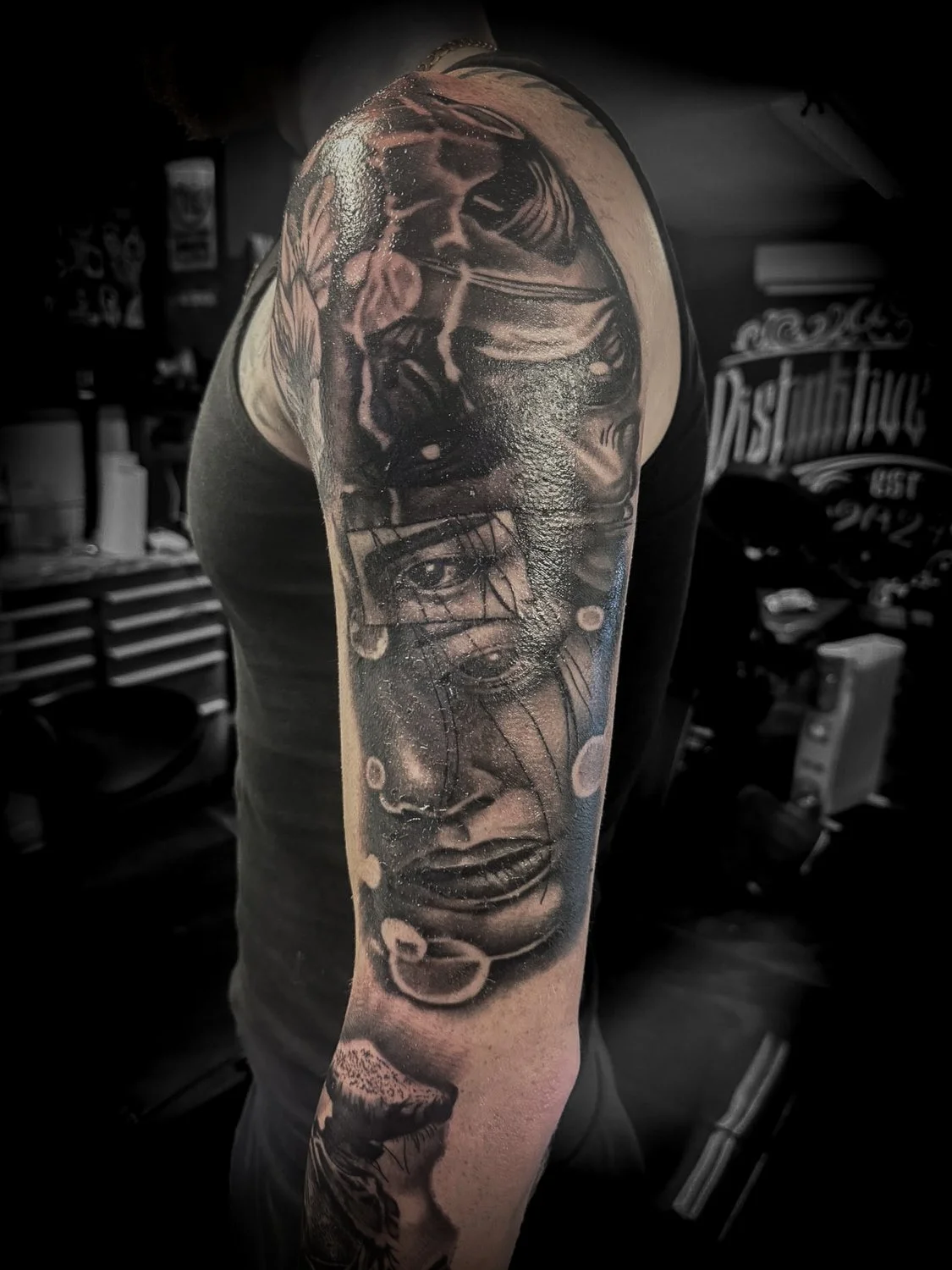 Black and gray tattoo sleeve featuring a woman's face, a face with sunglasses, and a spaceship, on an individual's upper arm in a tattoo studio.