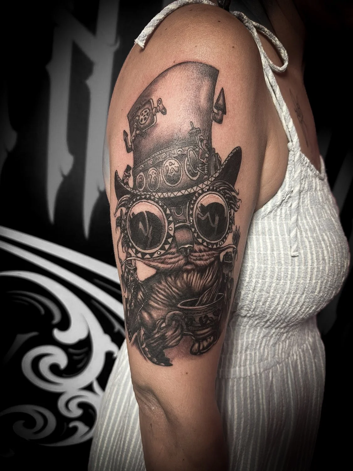 Black and gray tattoo of a cat wearing sunglasses and a top hat on a person's upper arm.