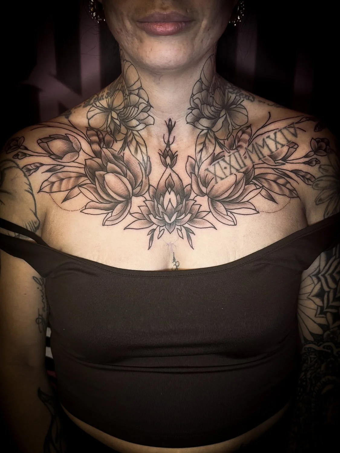 Close-up of a woman with tattoos on her chest, shoulders, and arms, wearing a dark off-shoulder top, with part of her face visible.