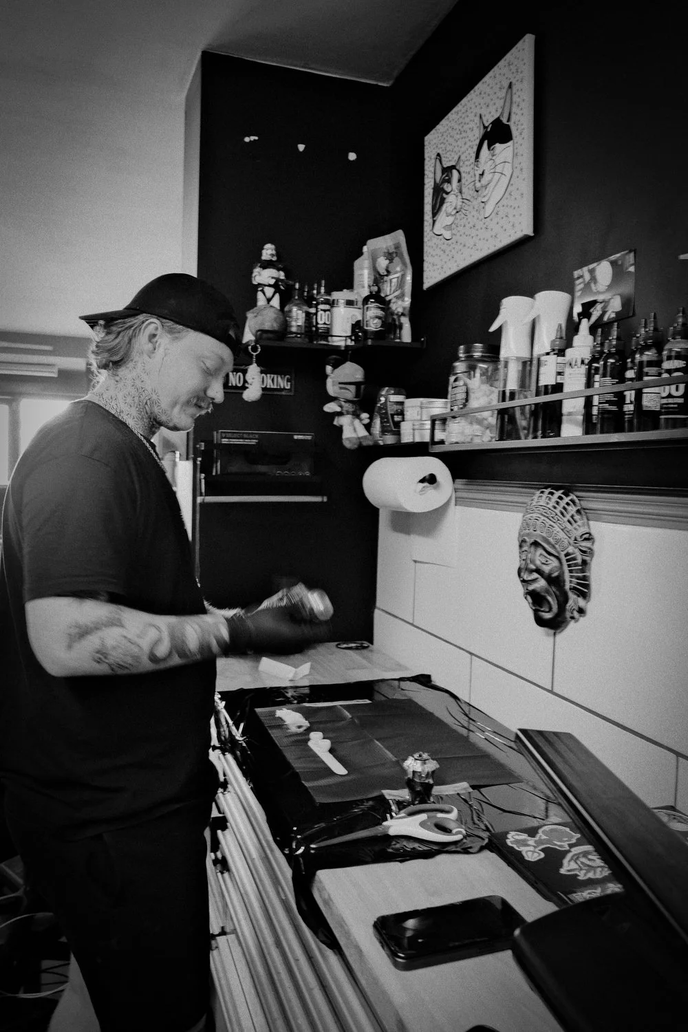 A tattooed man in a black cap and shirt working at a tattoo station in a tattoo parlor filled with art and supplies.