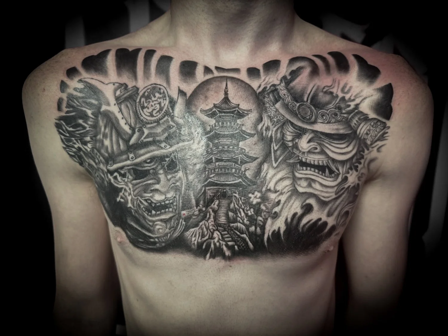 Black and gray tattoo of two Japanese masks, a pagoda, and a mountain scene on a person's chest.