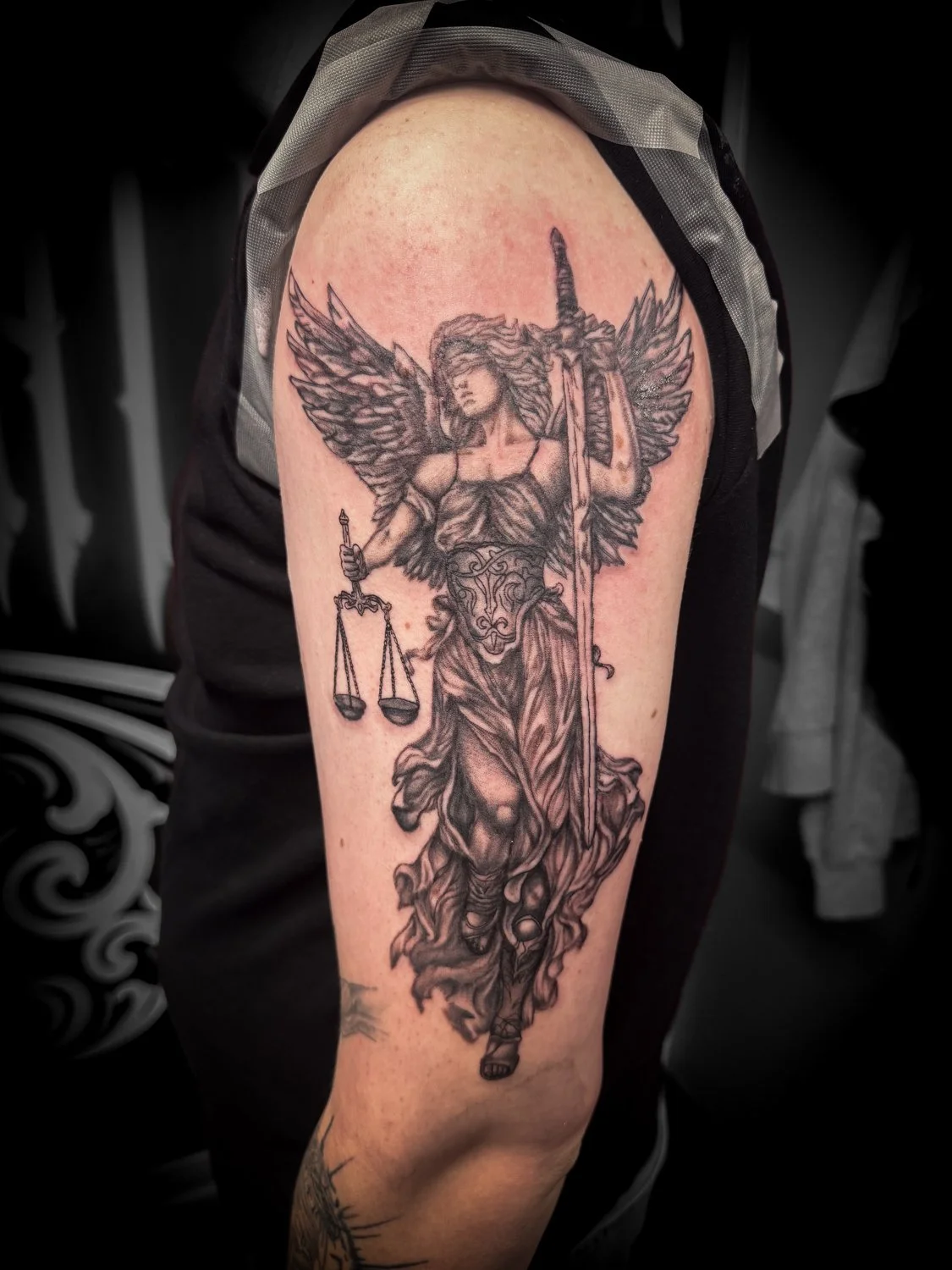 Black and gray tattoo of a winged woman holding a sword and a scale on the upper arm.