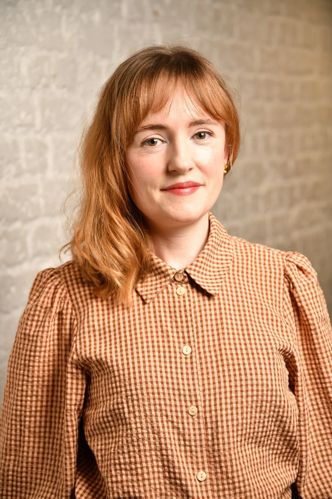 A woman with shoulder-length red hair, wearing a brown checkered blouse with a collar, standing against a textured beige wall.