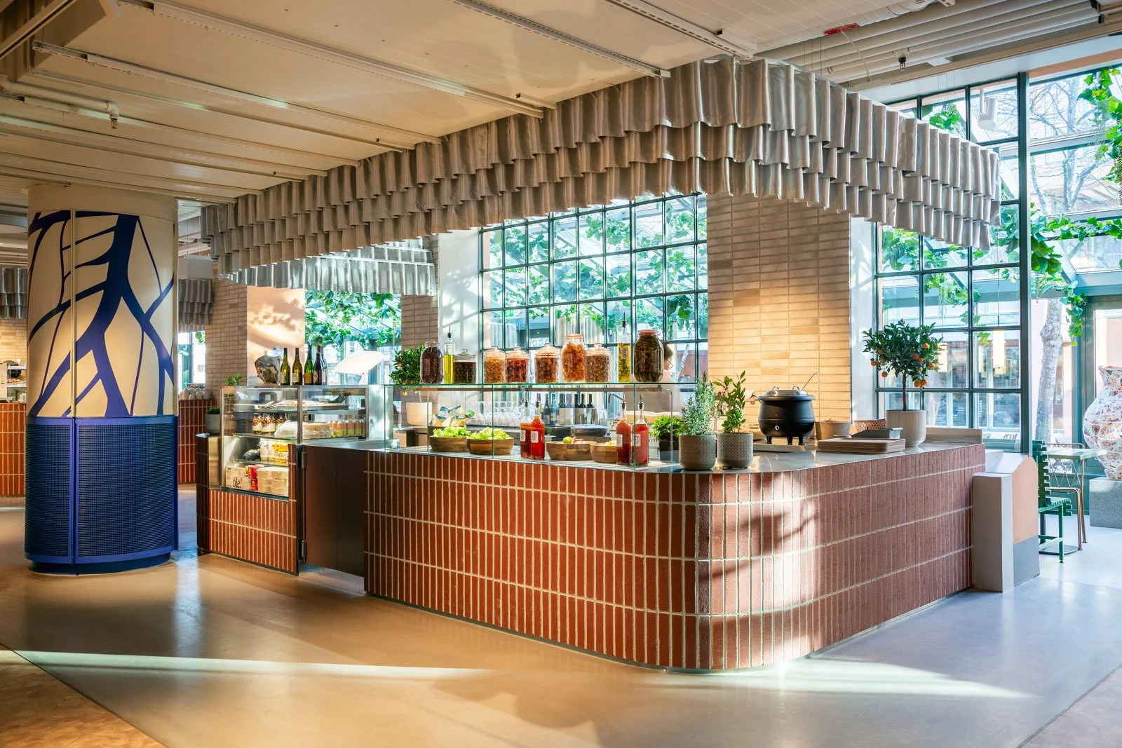 Ett modernt café med en rund bar med röda tegelstenar, glasdörrar för mat och dryck, flera växter, stora fönster som släpper in ljus, och takdekoration med rullande element i projektet Fabriken i Stockholm