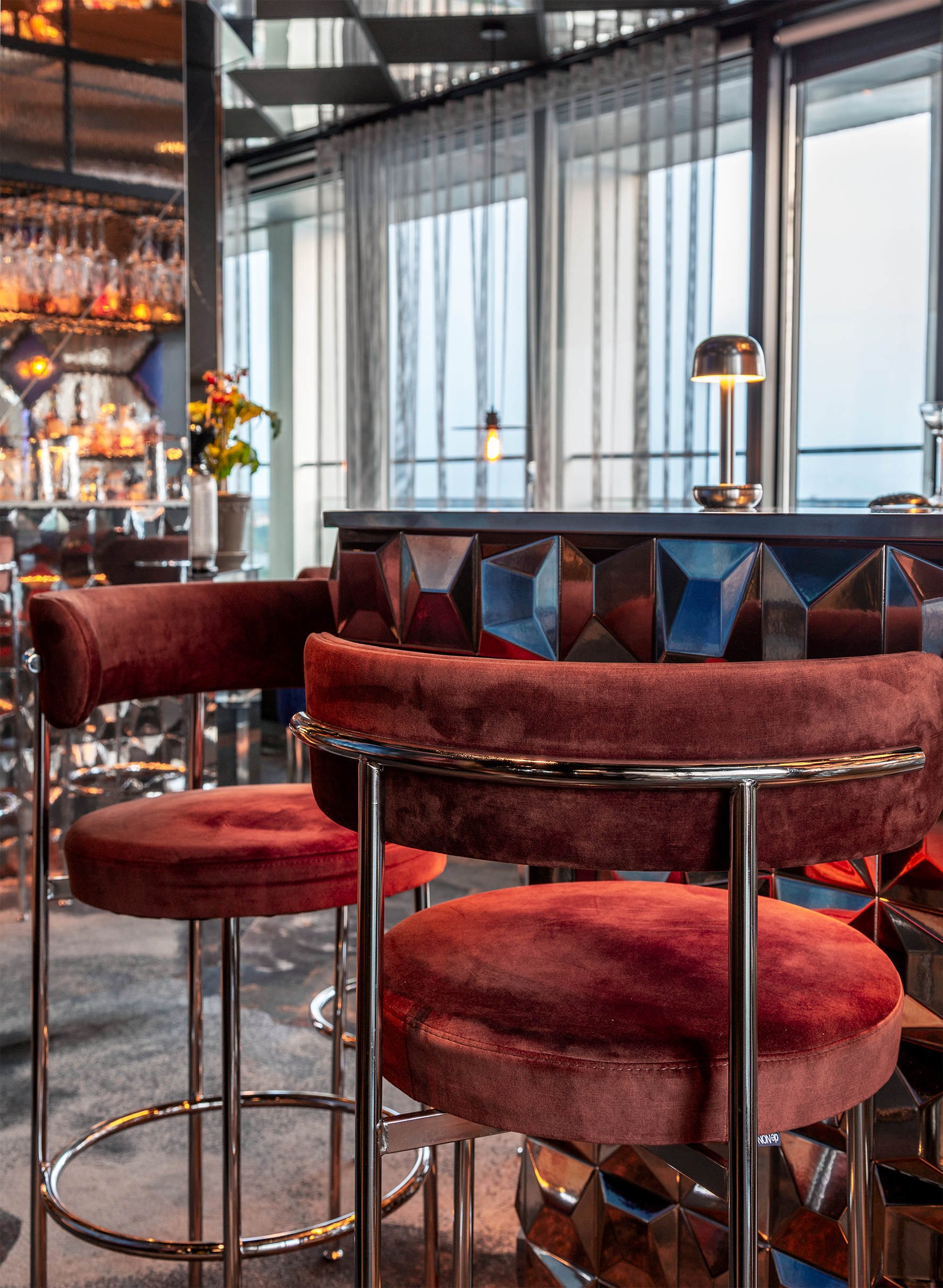 Detta är en bild av en modern bar eller restauranginteriör med röda sammetsstolar runt ett högt bord av metall och ett skåp med glas och flaskor i bakgrunden i projektet Le bar i Stockholm