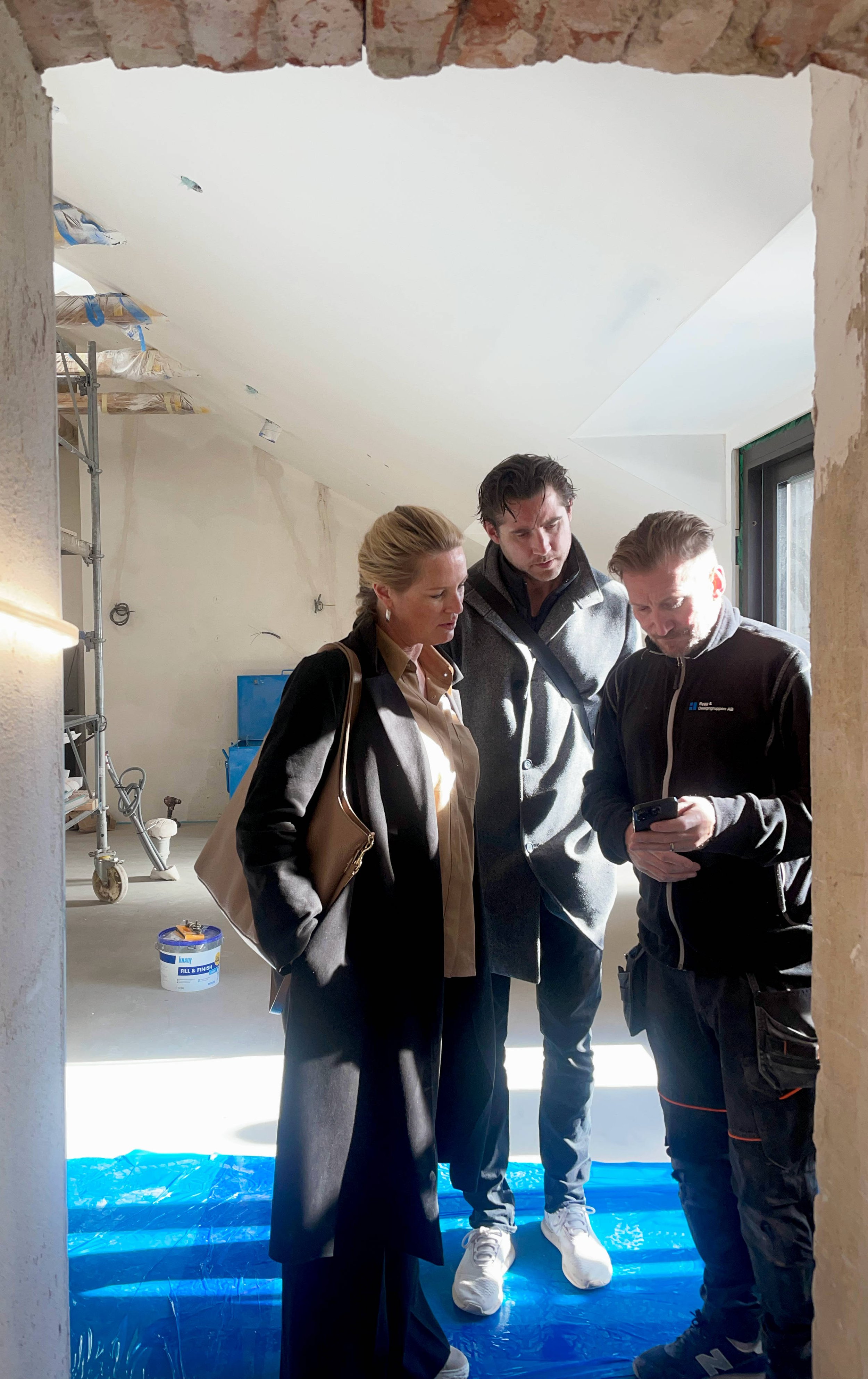 Tre personer står i ett rum under renovering, de tittar på en mobiltelefon. Rummet har byggnadsställningar, väggar under ombyggnad och en blå skyddsduk på golvet projektet i Skeppargatan 60 i Stockholm