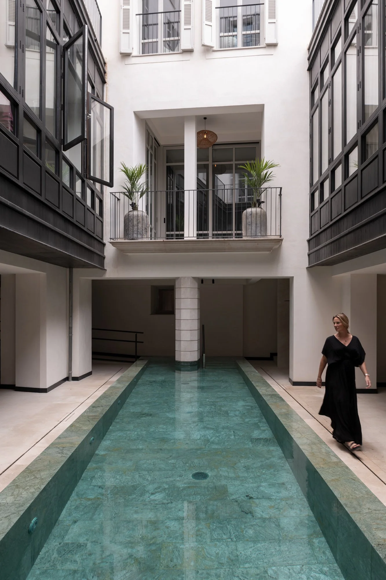 Inomhusgård med en pool eller vattenfunktion i mitten, omgiven av ljusa väggar och stora fönster. Det finns två våningar med balkonger och fönster, samt några gröna växter på balkongen i projektet Impremta Garden i Spain