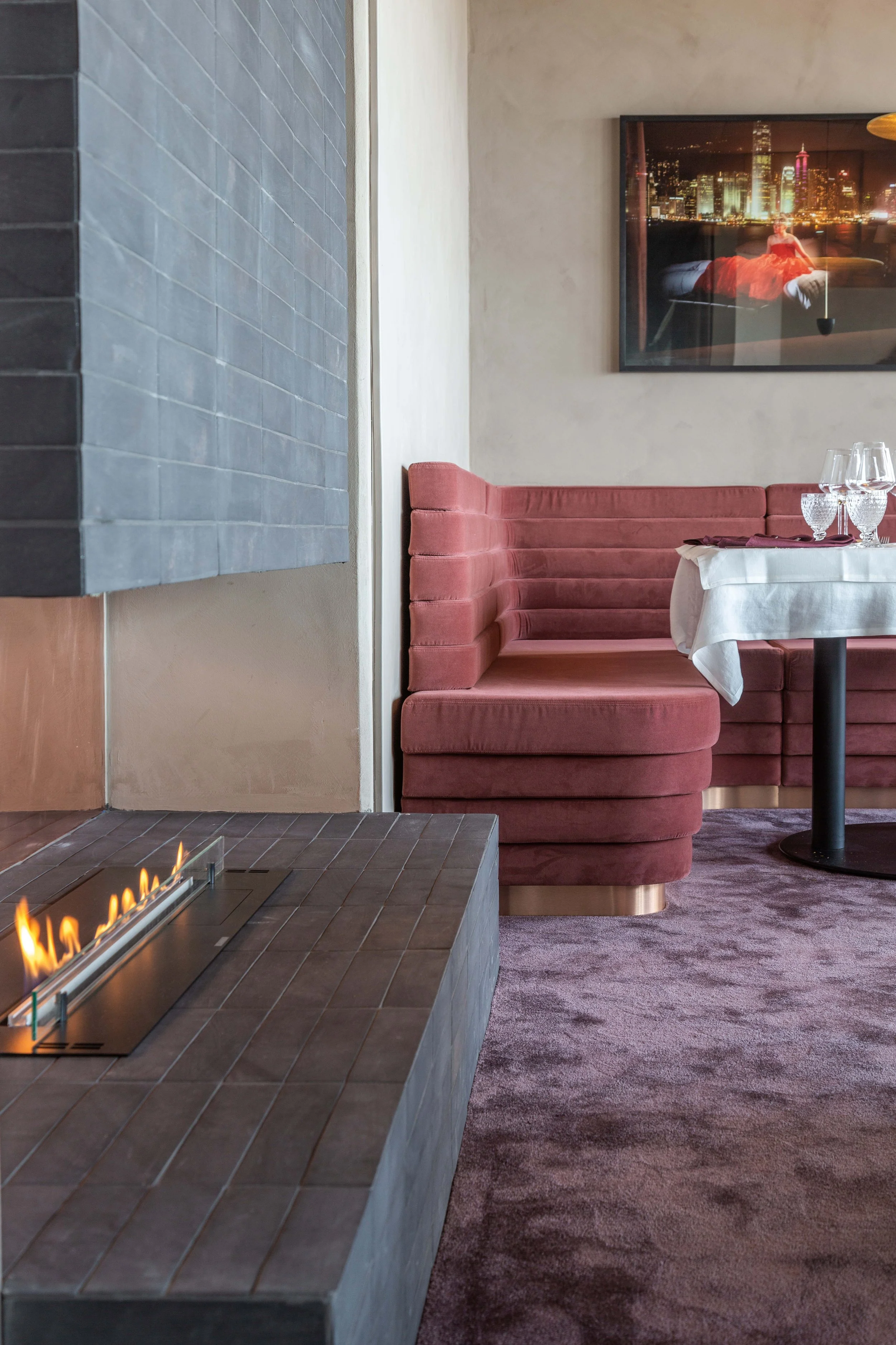 Inomhusbild av en restaurang eller ett kafé med en rosa soffhörna, ett runt bord med vita dukar och vin glas, ett stort konstverk på väggen samt en öppen spis. På bilden syns en stadssilhuett på konstverket i projektet La Tour i Stockholm
