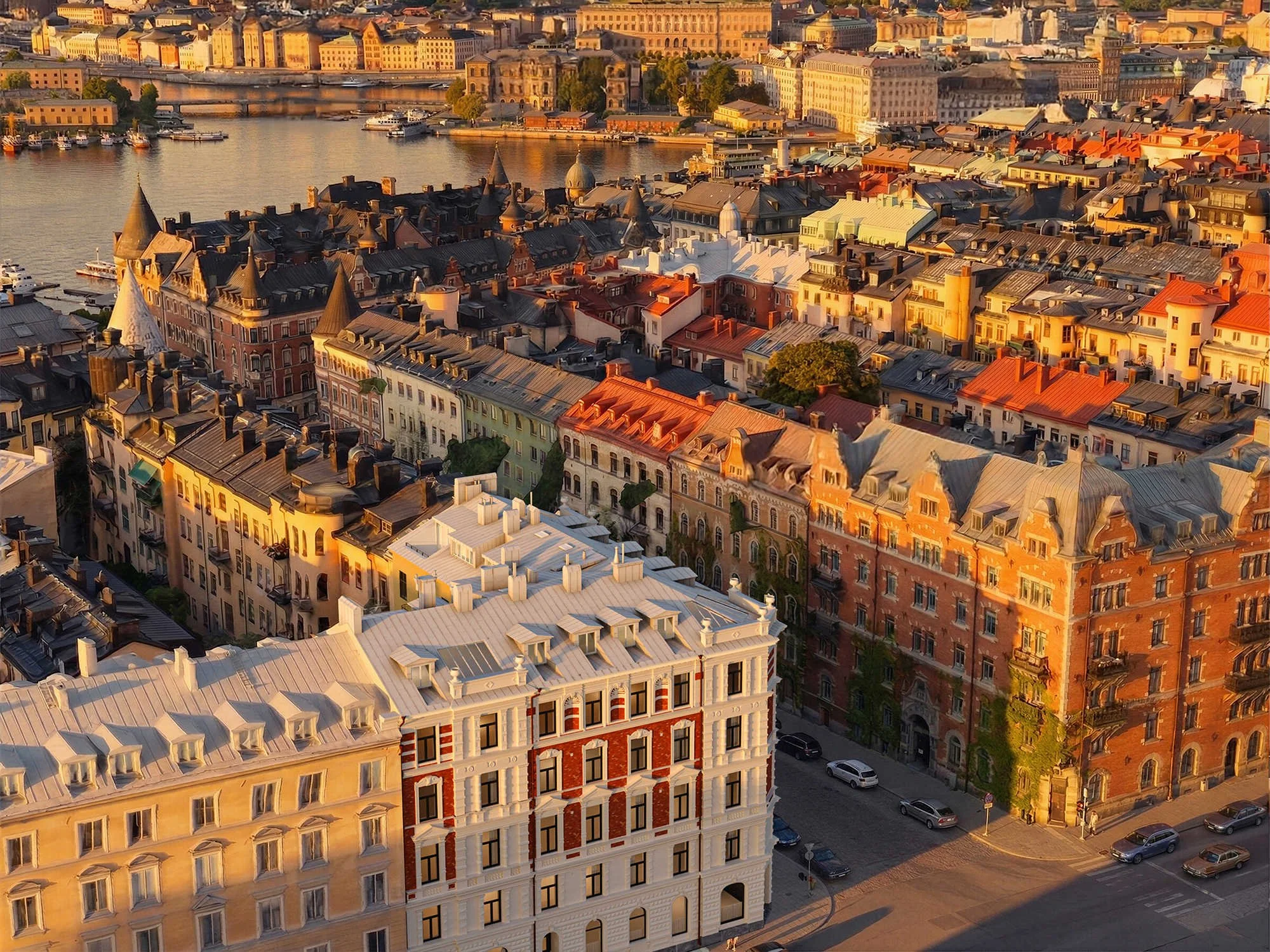 Vy över en stad vid vattnet vid solnedgång, med många byggnader med olika tak, inklusive torn och kupoler, samt båtar i vattnet och grönområden på andra sidan hamnen i projektet Torstenssonsgatan 12 i Stockholm