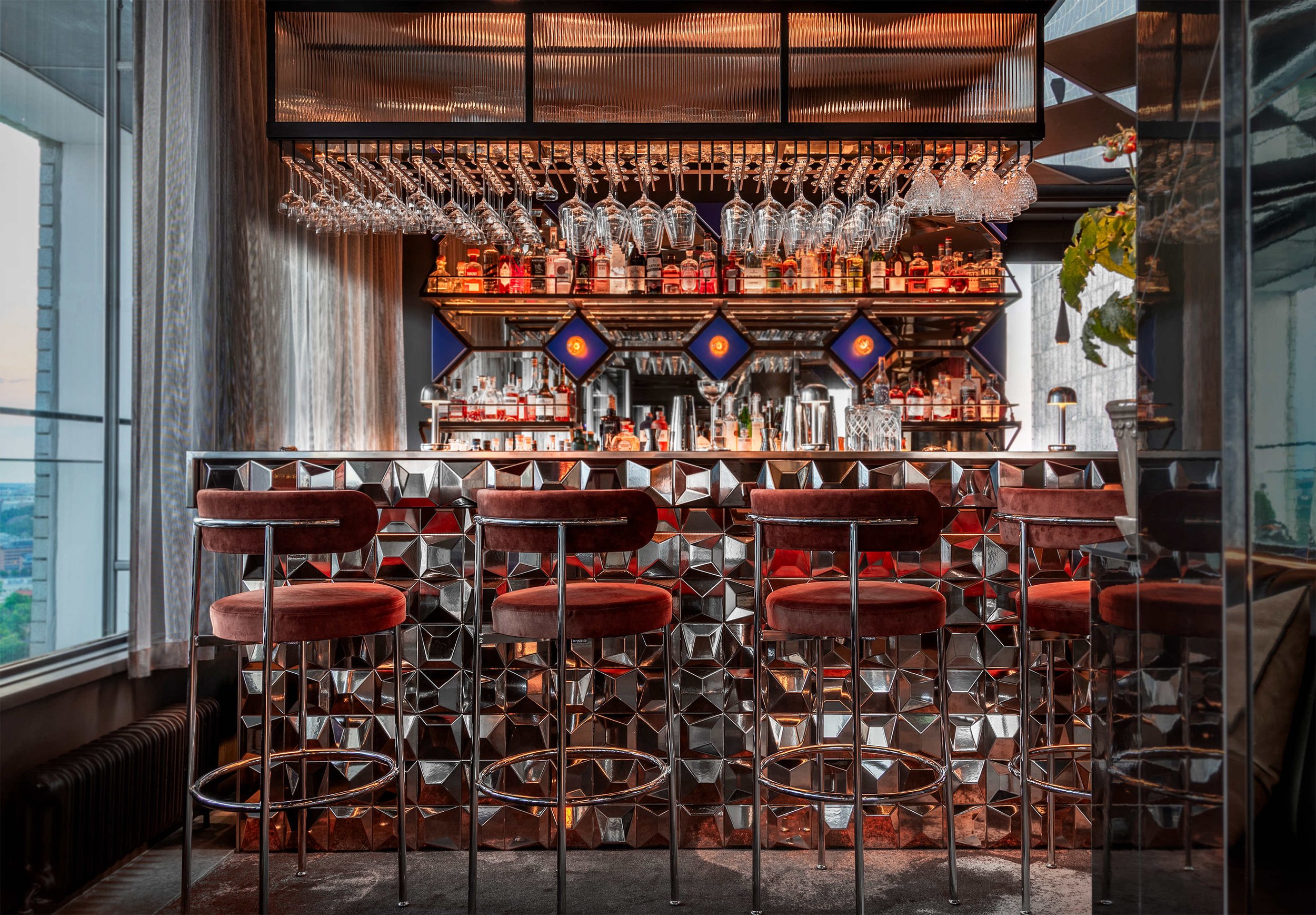En modern bar med glasögon och flaskor på hyllor, tre röda barstolar framför ett spegelvägg i projektet Le Bar i Stockholm