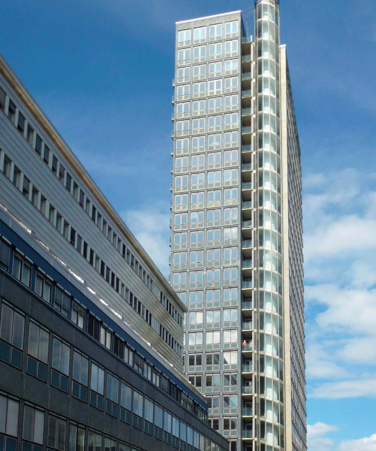 En hög modern kontorsbyggnad med speglande fönster och en annan byggnad i förgrunden, allt mot en klar blå himmel.