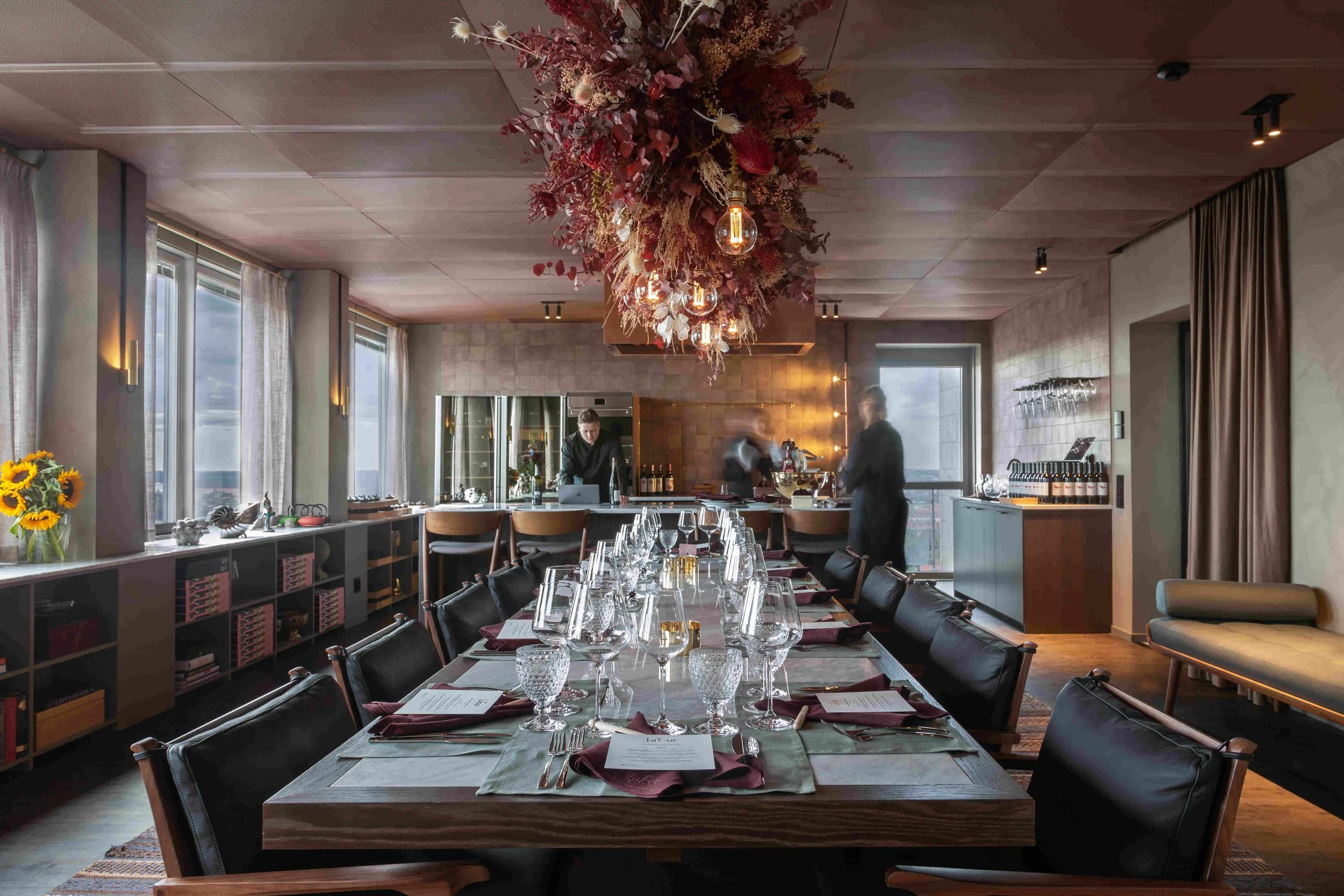 Ett elegant middagsrum med ett långbord som är dukat med glas, bestick, näsdukar och menyer för en formell middag, omgiven av svarta stolar och ett kök i bakgrunden i projektet La Tour i Stockholm