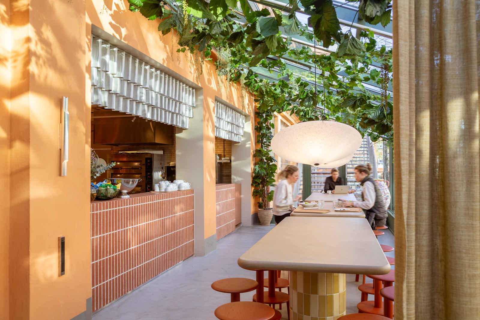 Interiör av ett modernt kafé med orange väggar, gröna växter och stora fönster. Det finns ett långt bord med bänkar och tre personer som arbetar eller fikar i projektet Fabriken i Stockholm