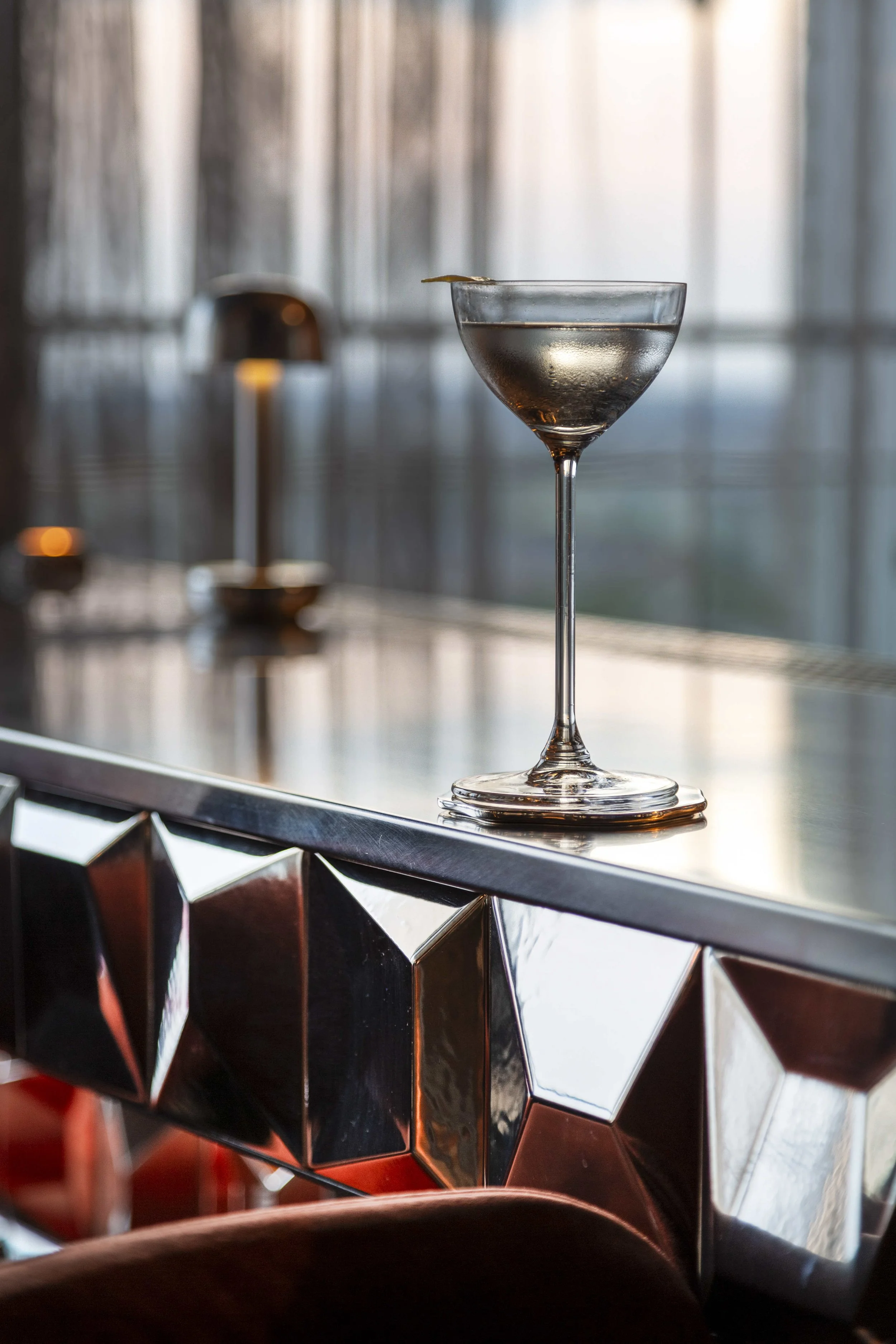 En cocktailskål utan dekoration placeras på en bardisk. Bakgrund av fönster med ljus silar genom, vilket skapar en ljus och lugn atmosfär i projektet Le Bar i Stockholm