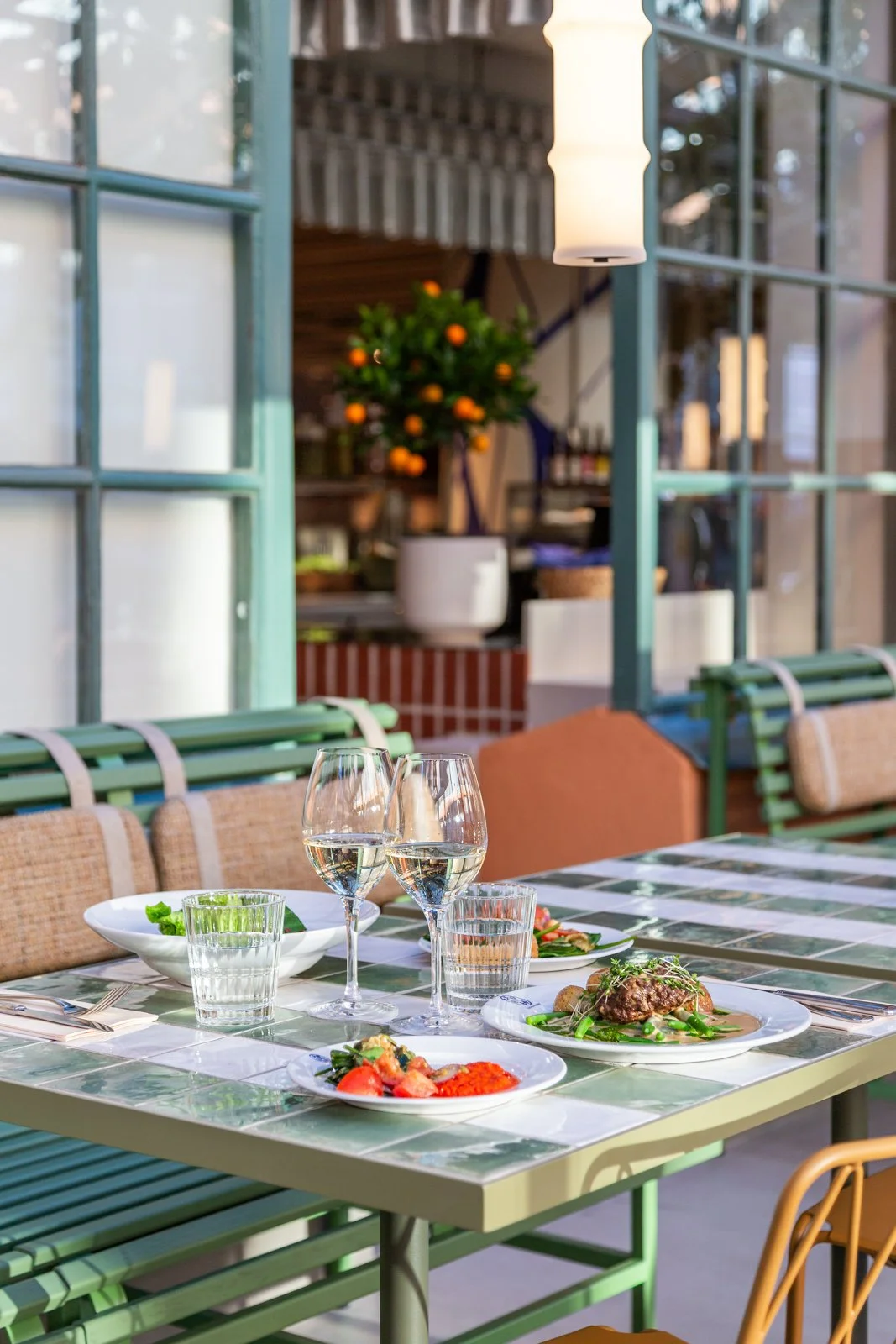 Matbord med tallrikar, glas med vin, vatten och mat, i ett uterum med gröna möbler, blommande växt och glasdörrar i projektet Fabriken i Stockholm