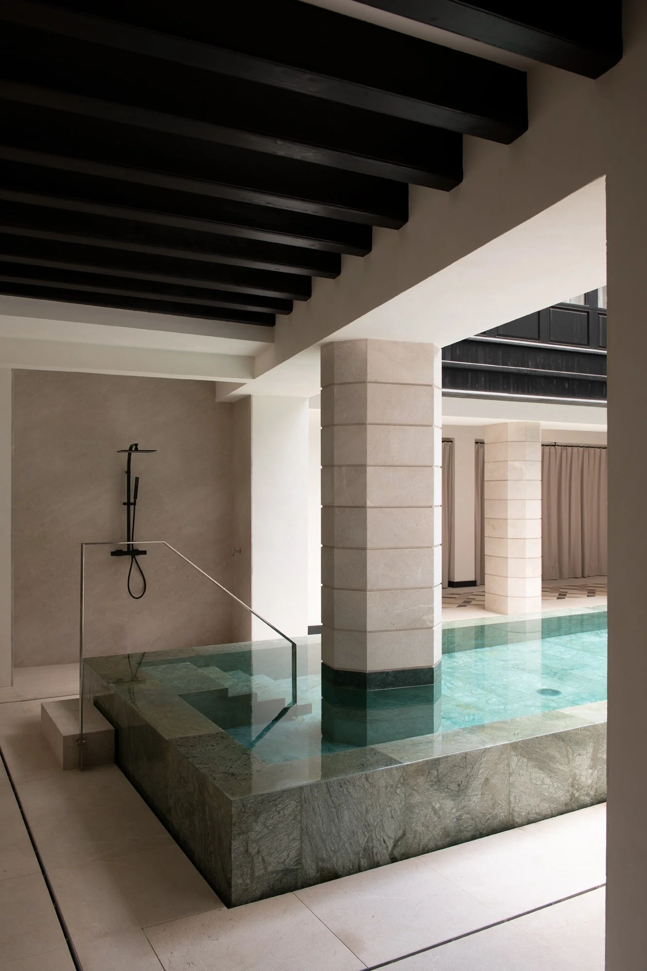 Inomhuspool med ett väggduschområde med svart duscharmatur, omgiven av modern arkitektur med vita väggar, en pool av grönt marmor och pelare, samt ett område med gardiner i bakgrunden i projektet Impremta garden i Spain