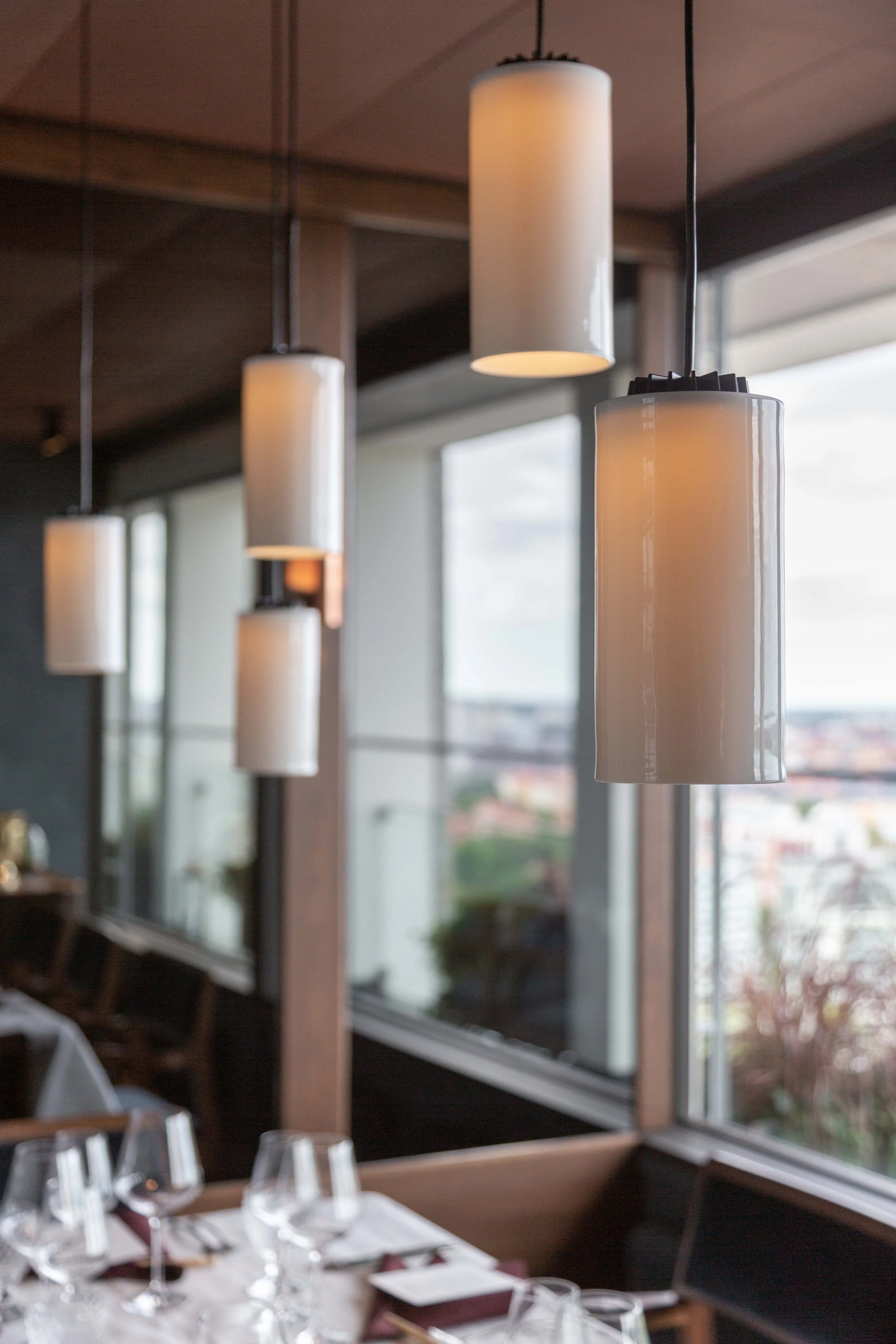 Tiered white pendant lights hanging from a wooden ceiling in a restaurant, with a large window in the background showing an urban cityscape i projektet La Tour i Stockholm